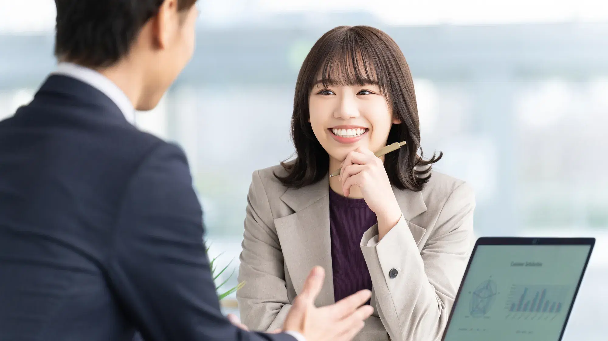 灰色のスーツを羽織っている女性とパソコンを置きながら女性と会話している紺色のスーツを着た男性