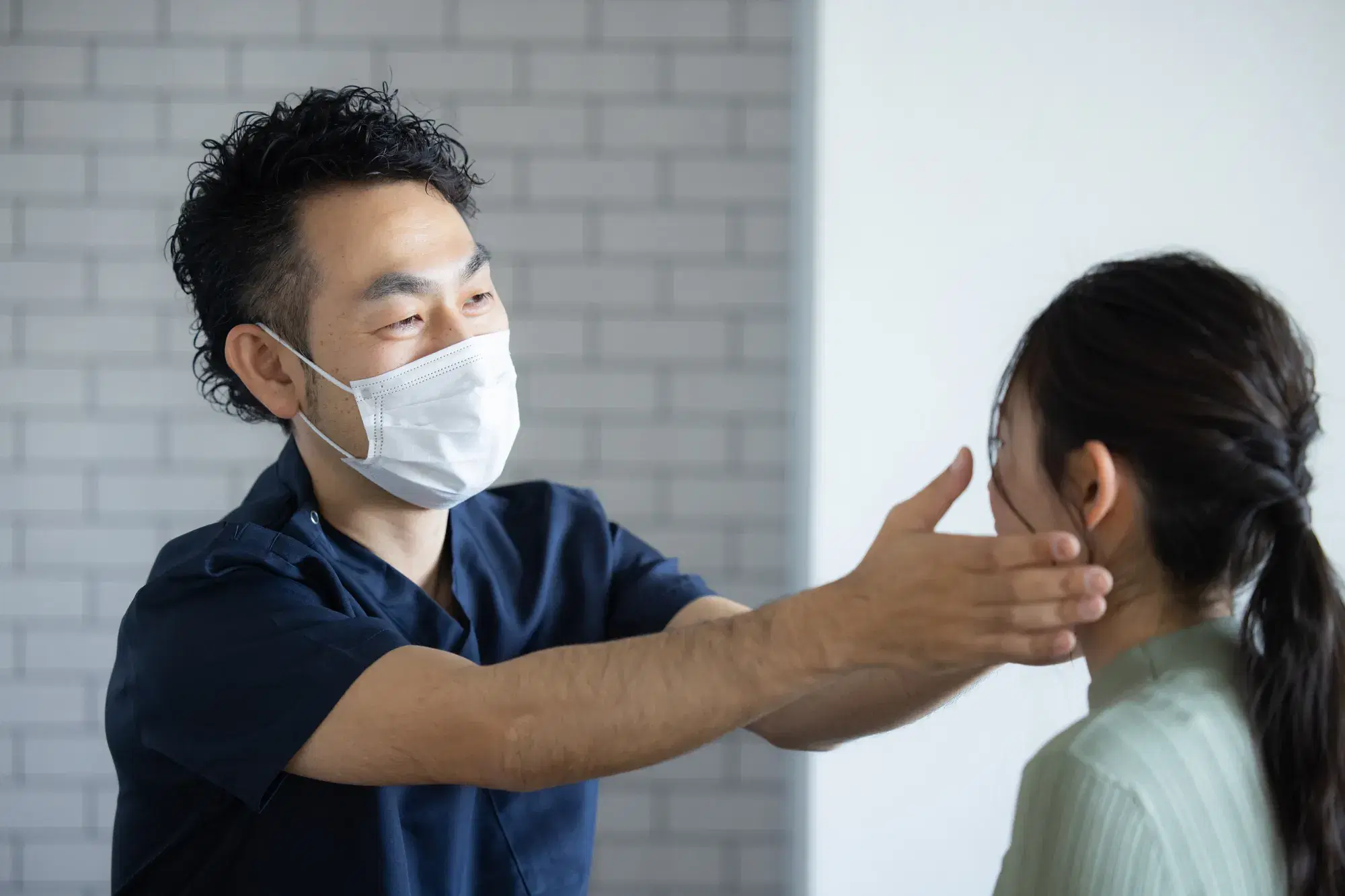 髪の長い女性の耳元を手で触って検診する青い服を着たマスクの医者