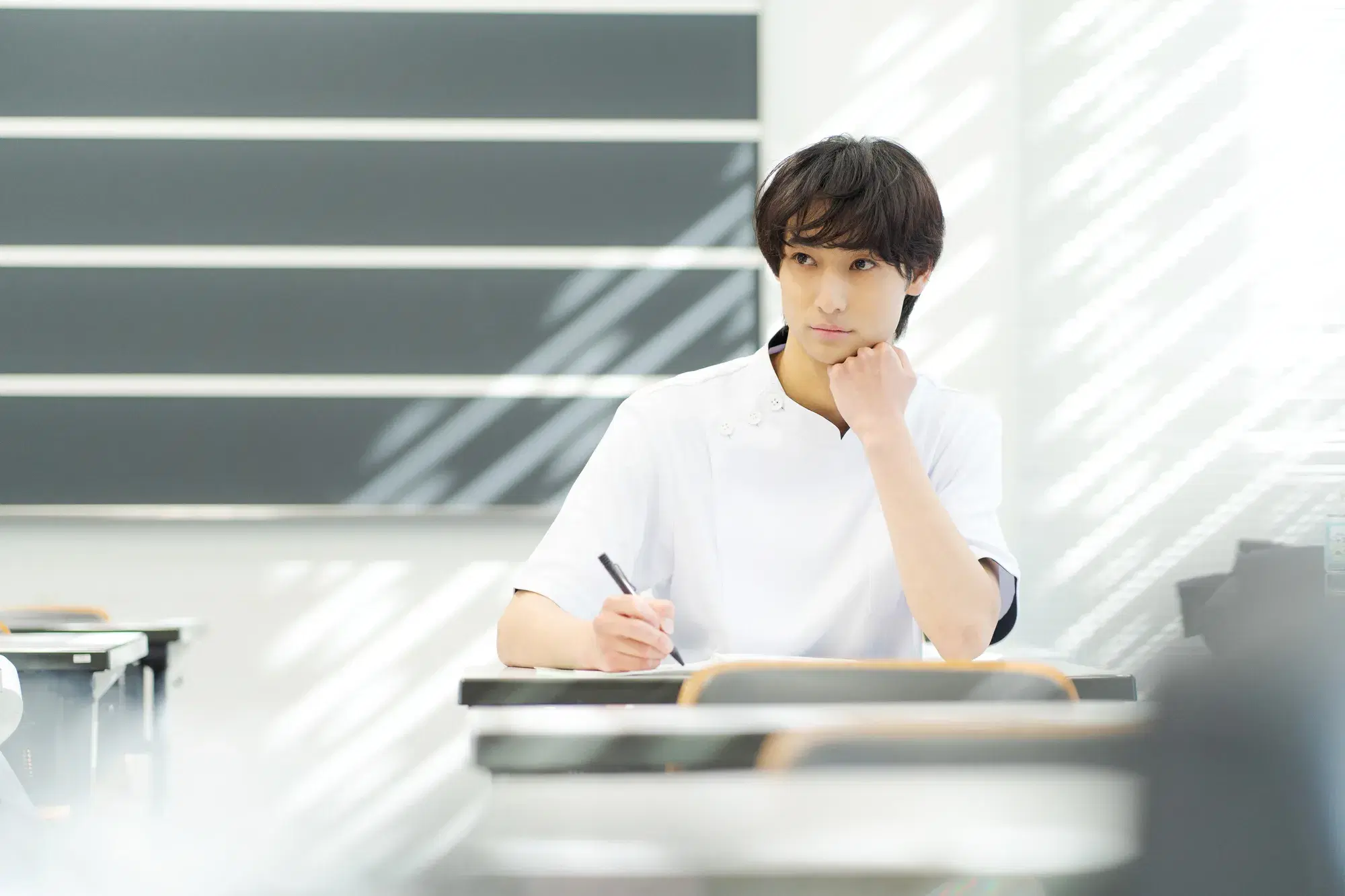 白い服を着た男性が講義室の一番後ろの席で顎に手を着ながら勉強している。