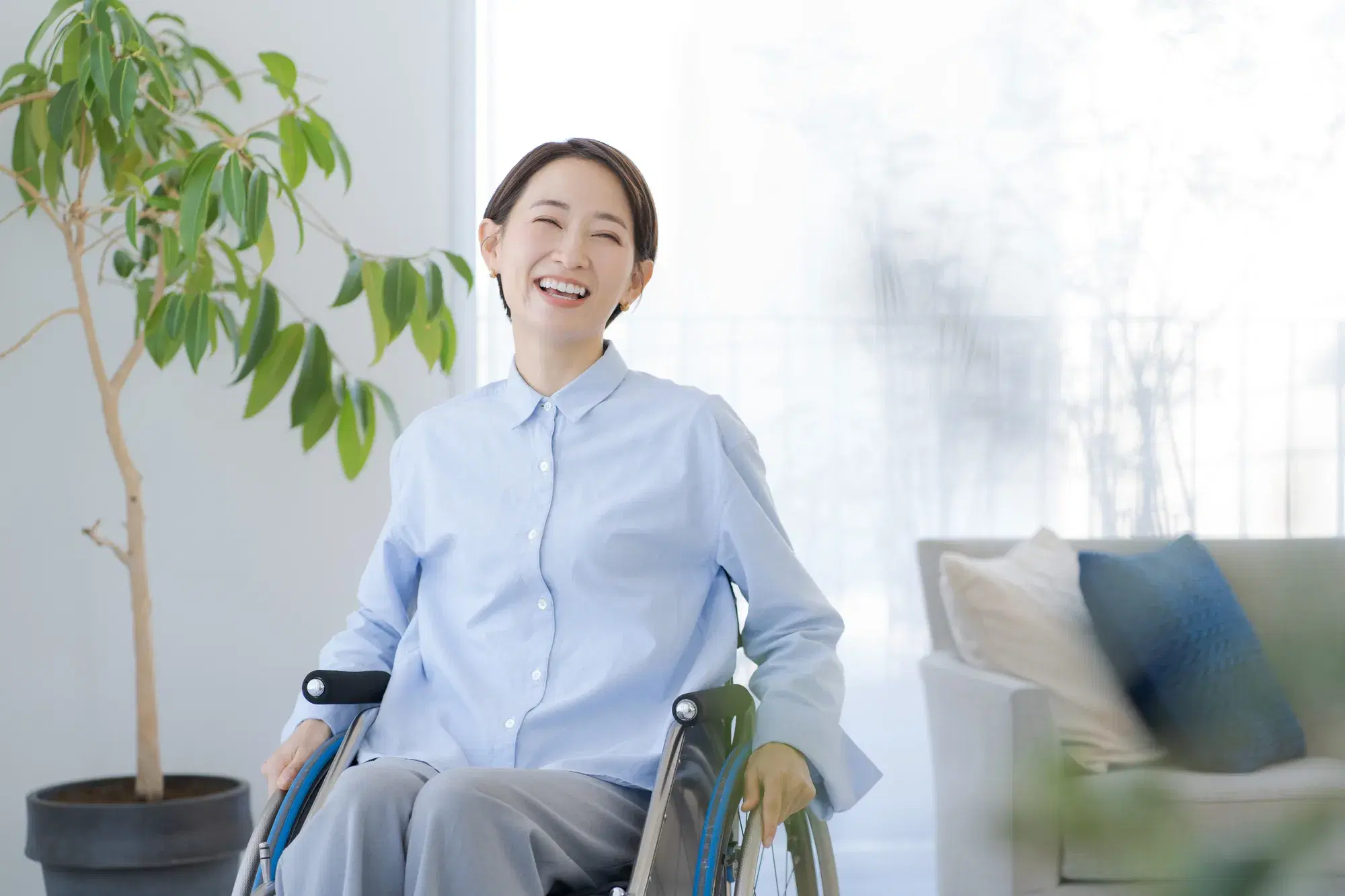 ソファーと観葉植物がある空間内で車椅子に座る水色の服を着た笑顔の女性