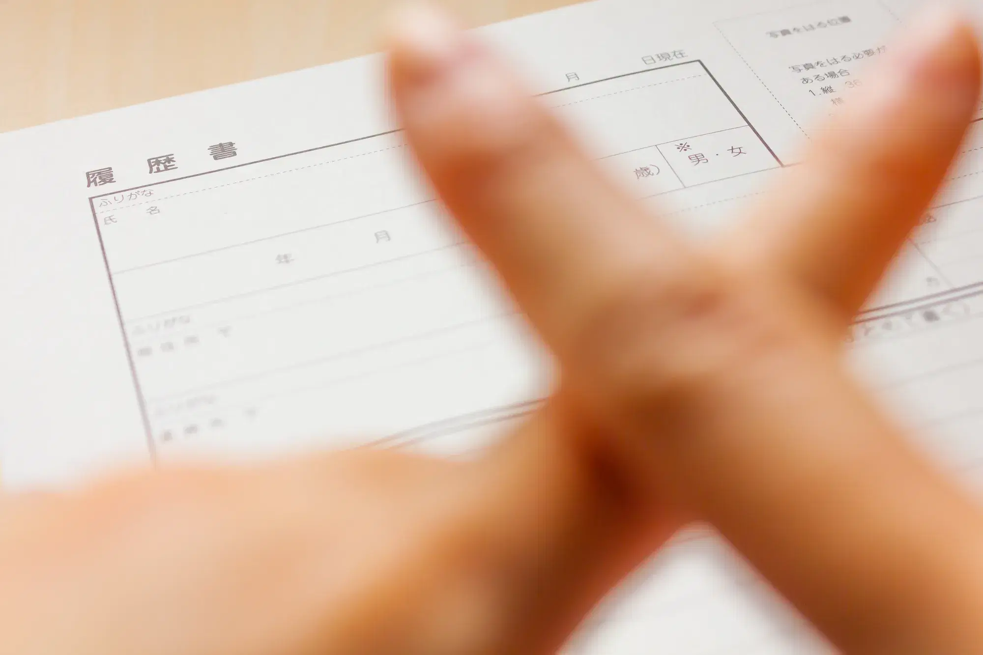 「履歴書」と書かれた用紙の手前で、指を交差させてバツ印を作っている手元が写っている。