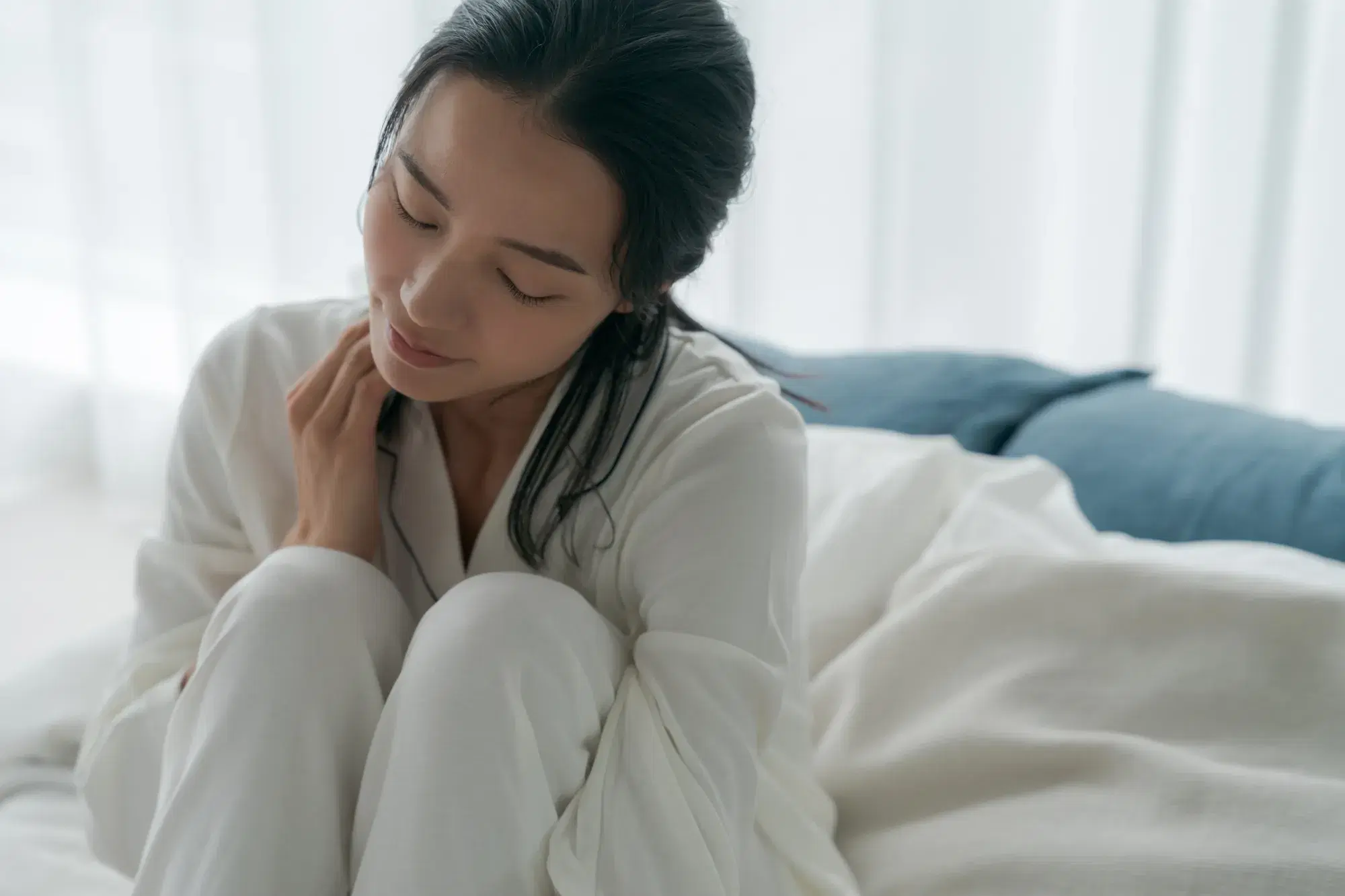白いパジャマを着た女性が、ベッドの上で膝を抱えて座っている。