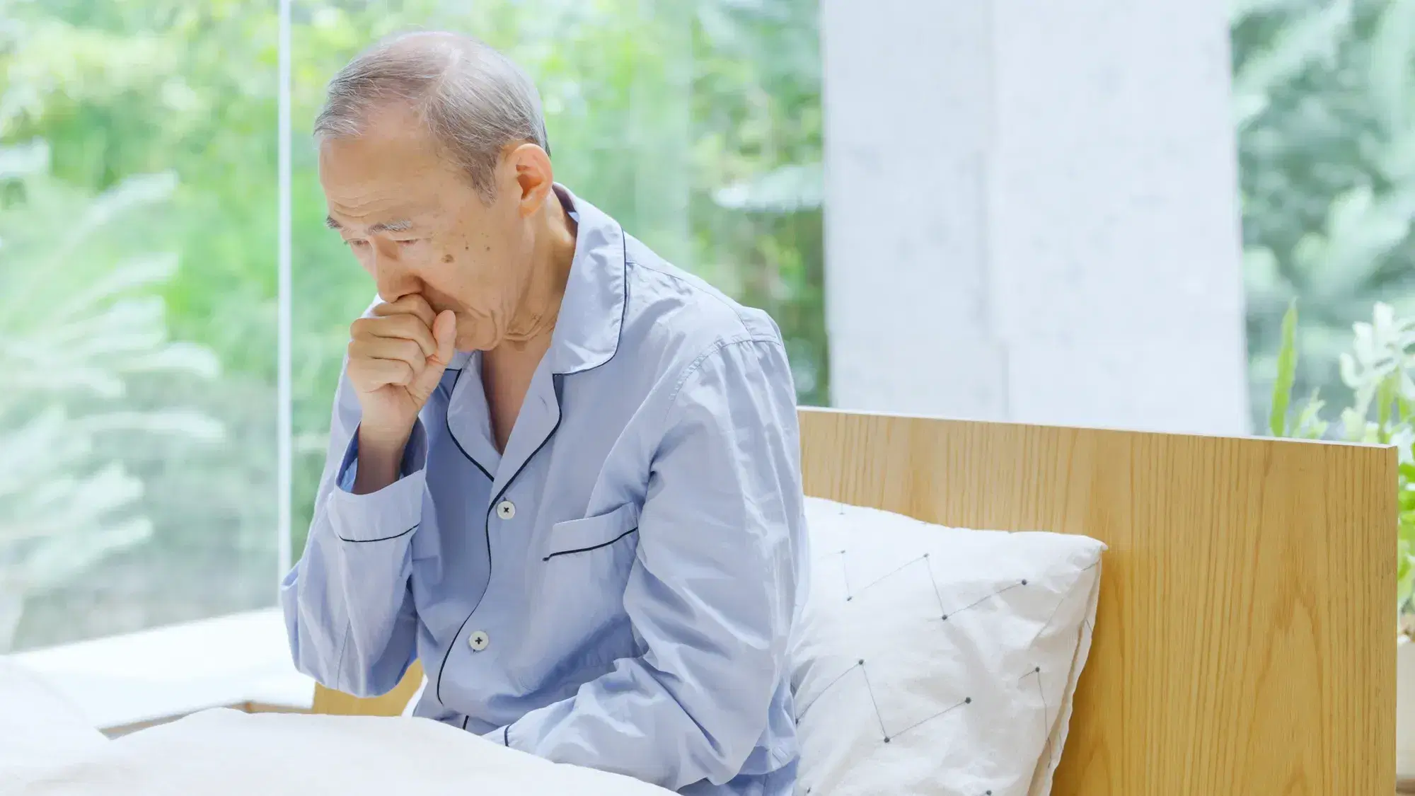 青い服を着た白髪の男性がベッドの上でせき込んでいる