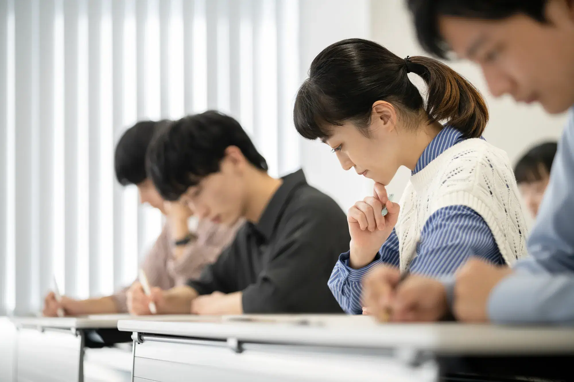 複数の人数の中で髪を結んだ女性が顎に手を当てながら考え込む