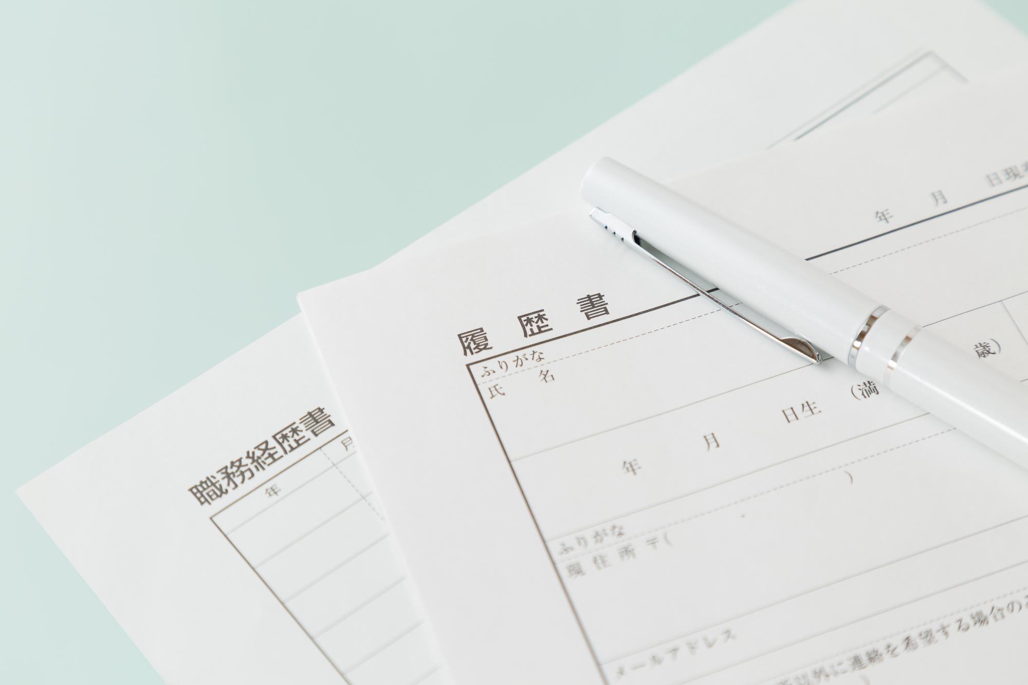 「履歴書」と「職務経歴書」と書かれた用紙の上に白いペンが置かれている様子が写っている。