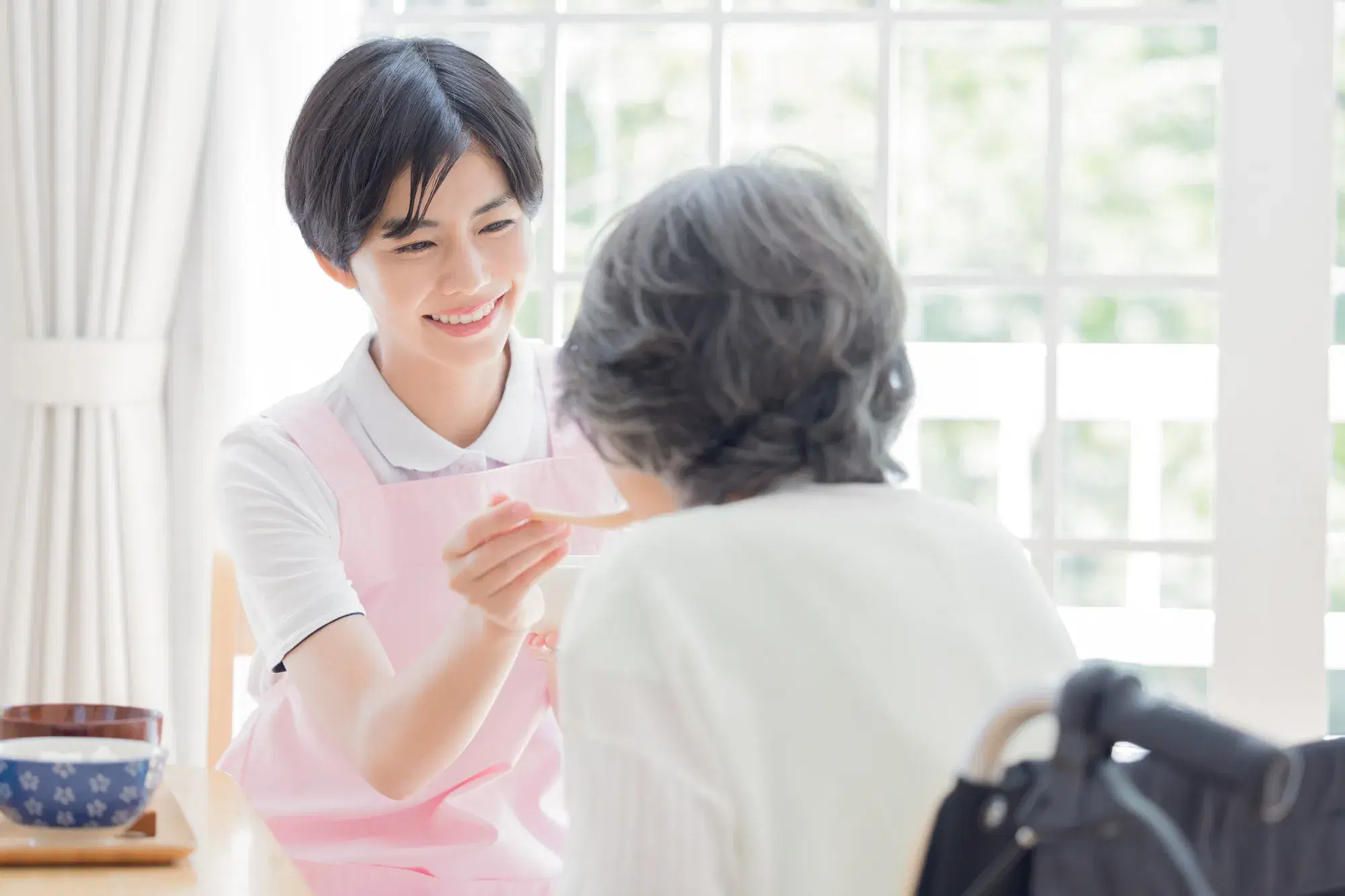 ピンク色のエプロンを着た女性が白色の服を着た年配の女性を介護