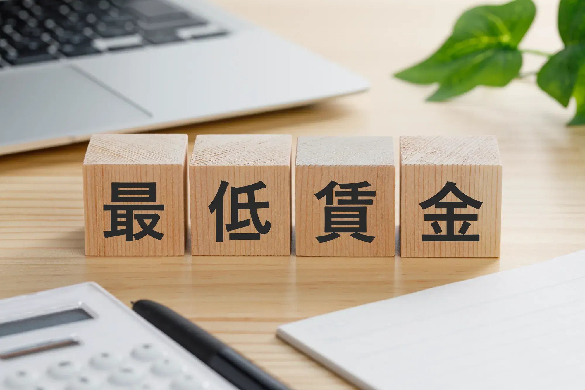 机の上に「最低賃金」と書かれた木製ブロック、ノートパソコン、電卓、用紙が置かれている様子が写っている。