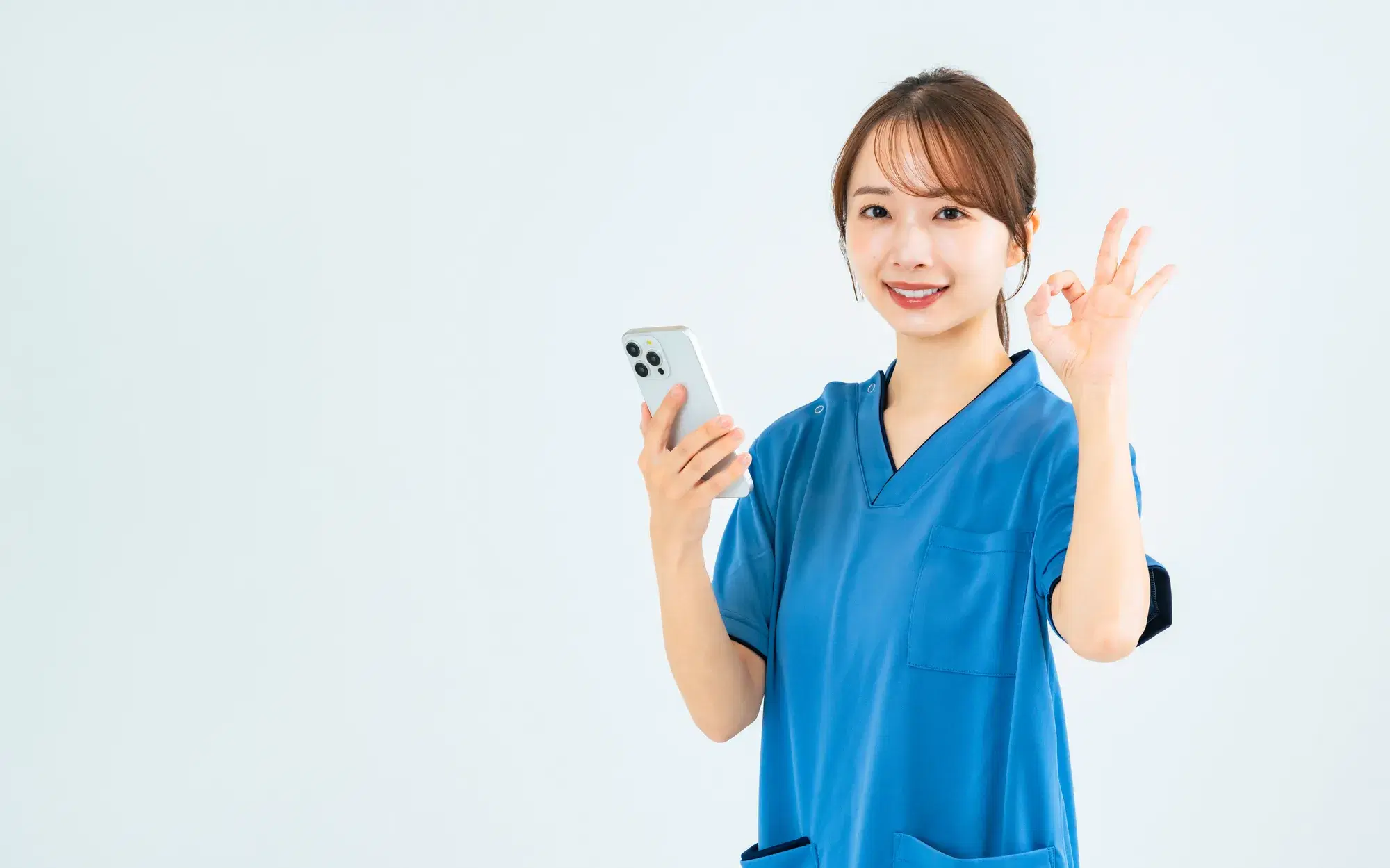 青い服を着た医療従事者が、右手にスマートフォンを持って左手でOKのマークを作っている。