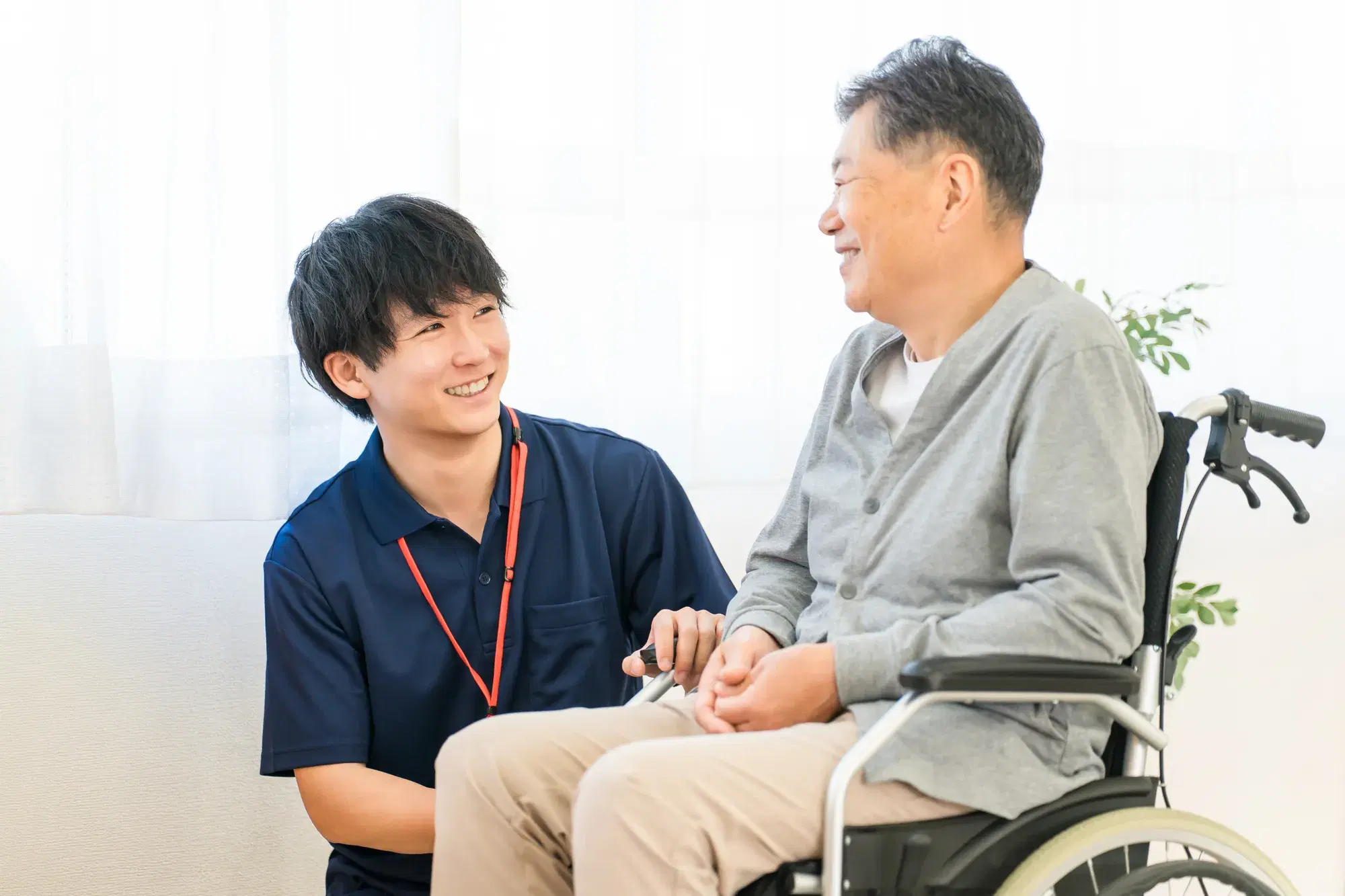 灰色の服を着た車椅子に座る年配の男性を紺色の服を着た男性が介護