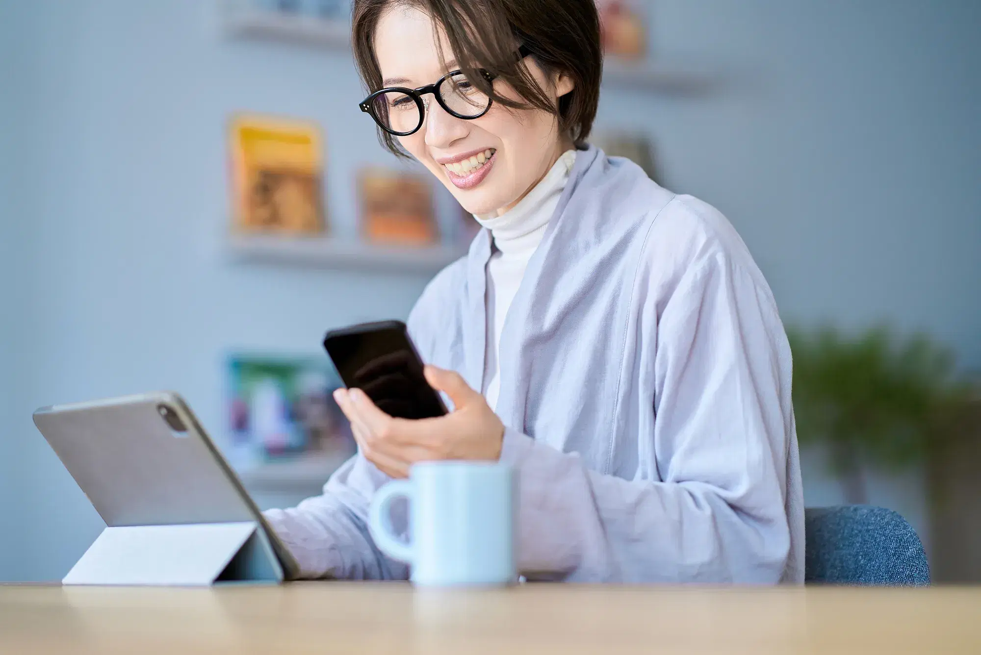 机の上に横向きで建てられたタブレットと左でスマホを操作する女性