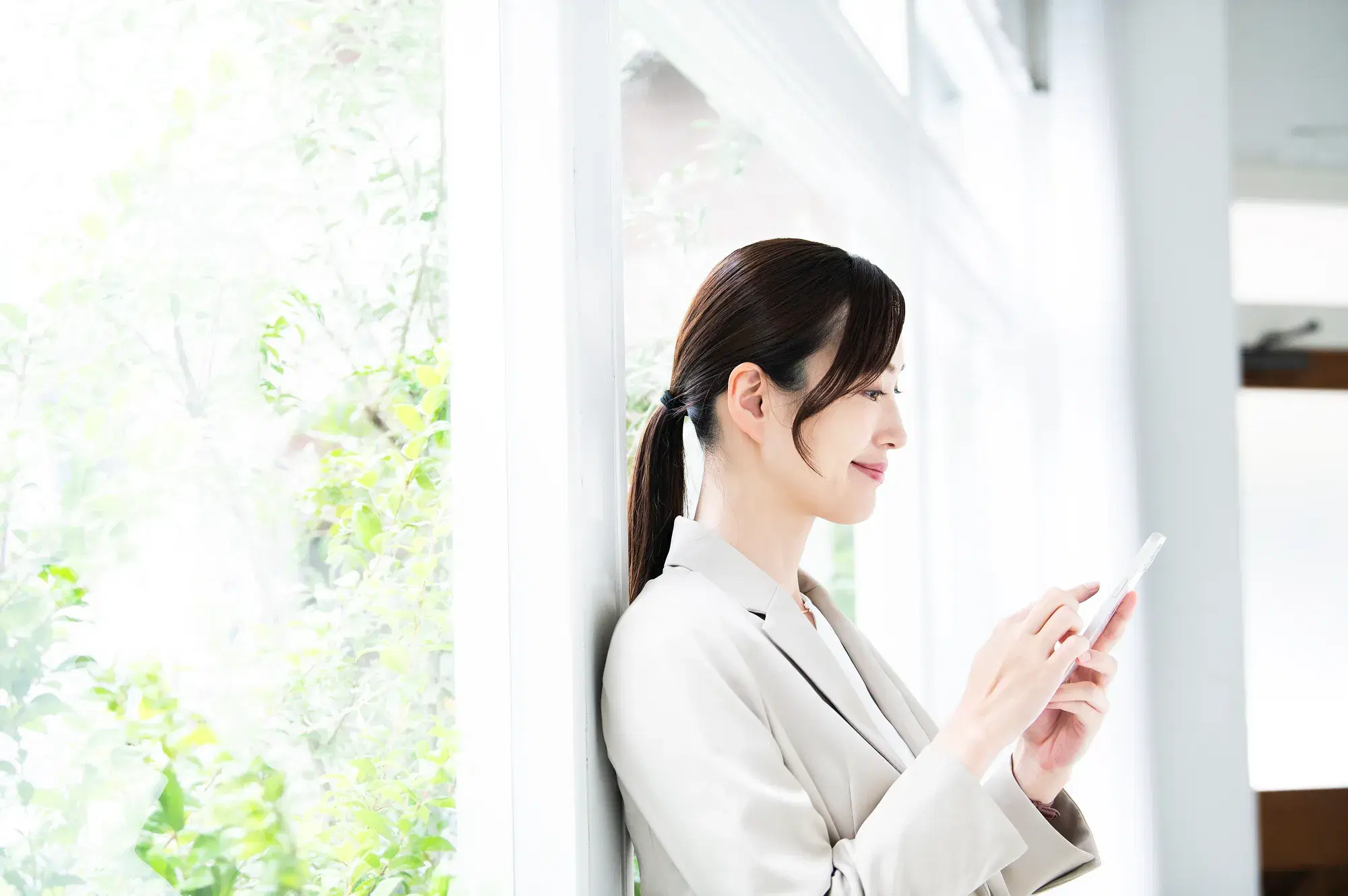 ガラス窓を囲む枠組みにもたれかかる白いスーツを着たスマホを操作する女性