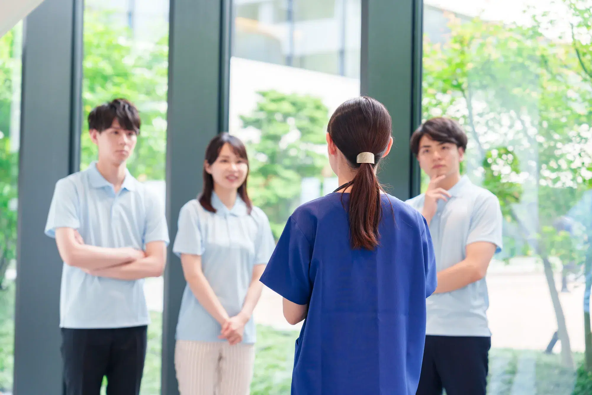 ガラス窓がある空間で手前に青色の服を着た女性と奥側に水色の服を着た男女3人