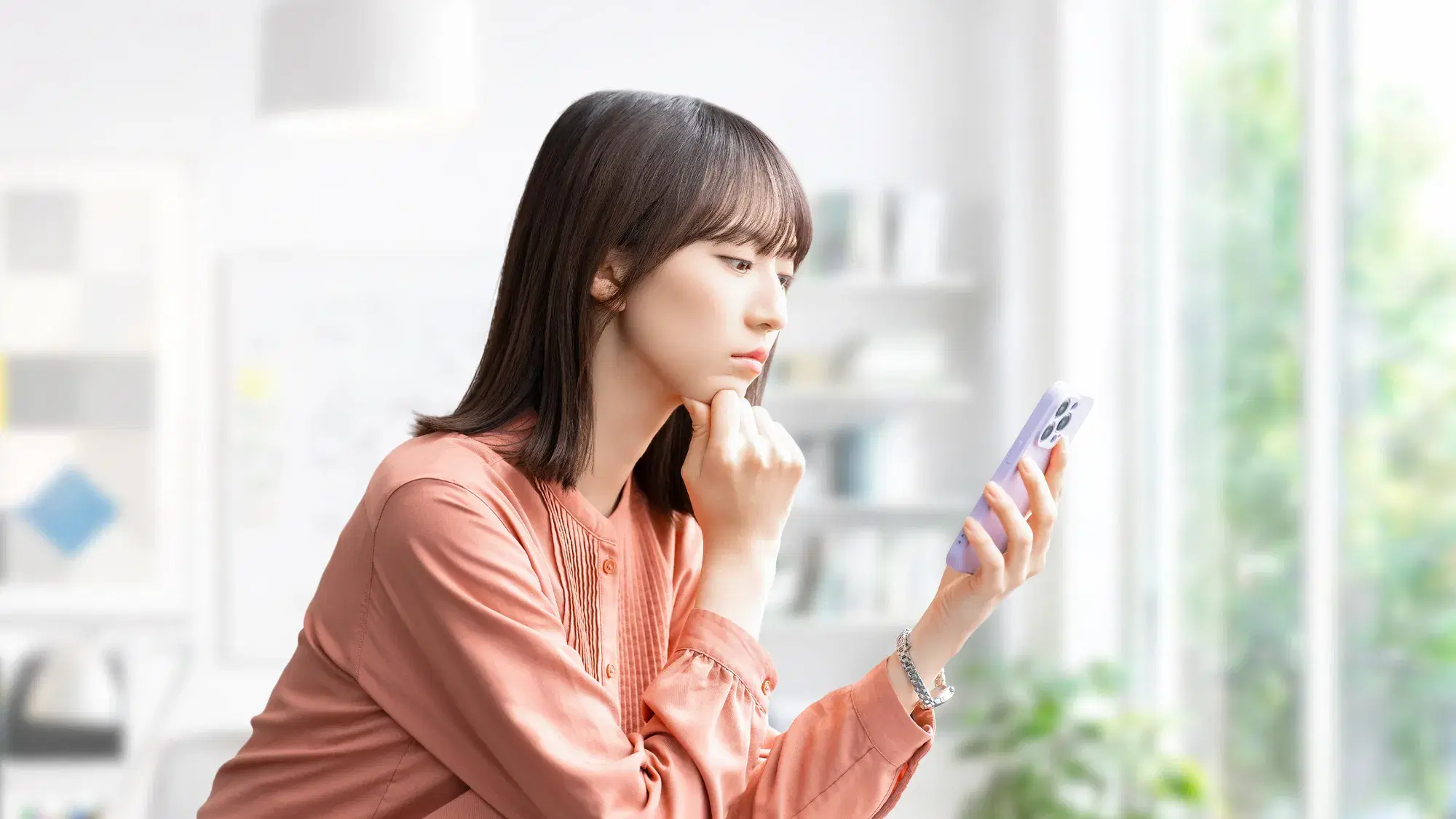 オレンジの服を着た若い女性が机に肘をつき、右手は顎につけ、左手に持ったスマートフォンを使用している