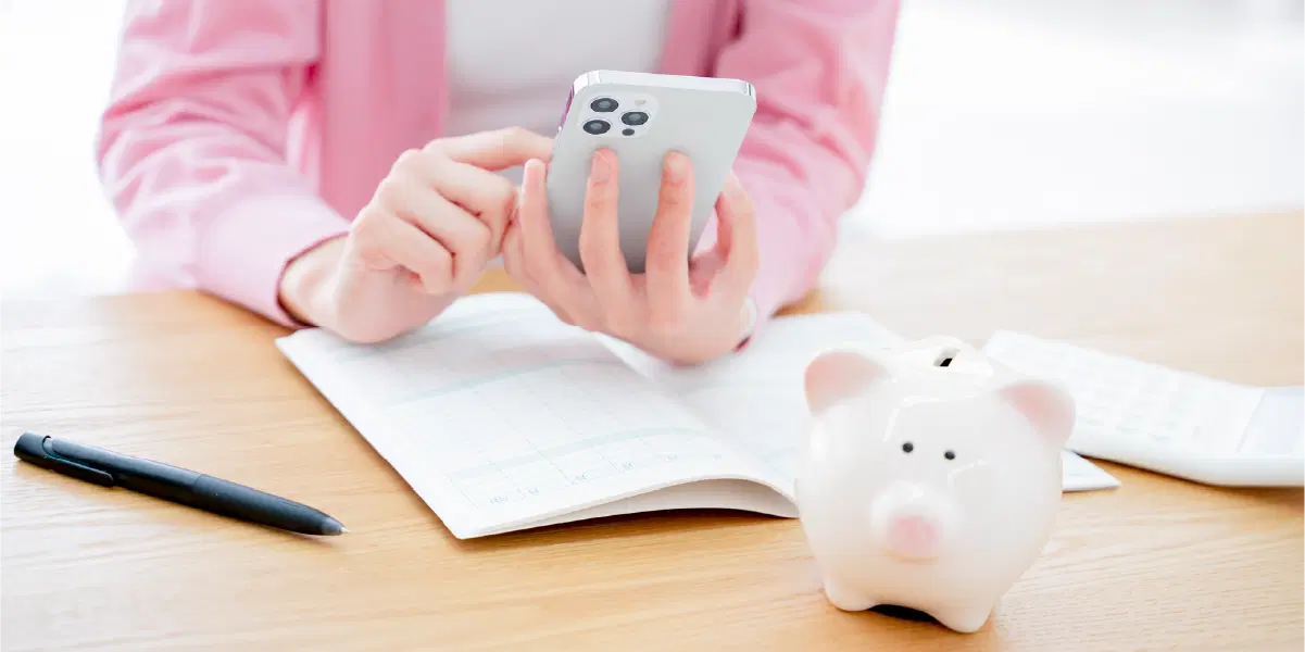 豚の貯金箱、開かれたノート、ペンが机の上に置かれ女性はスマホを操作している