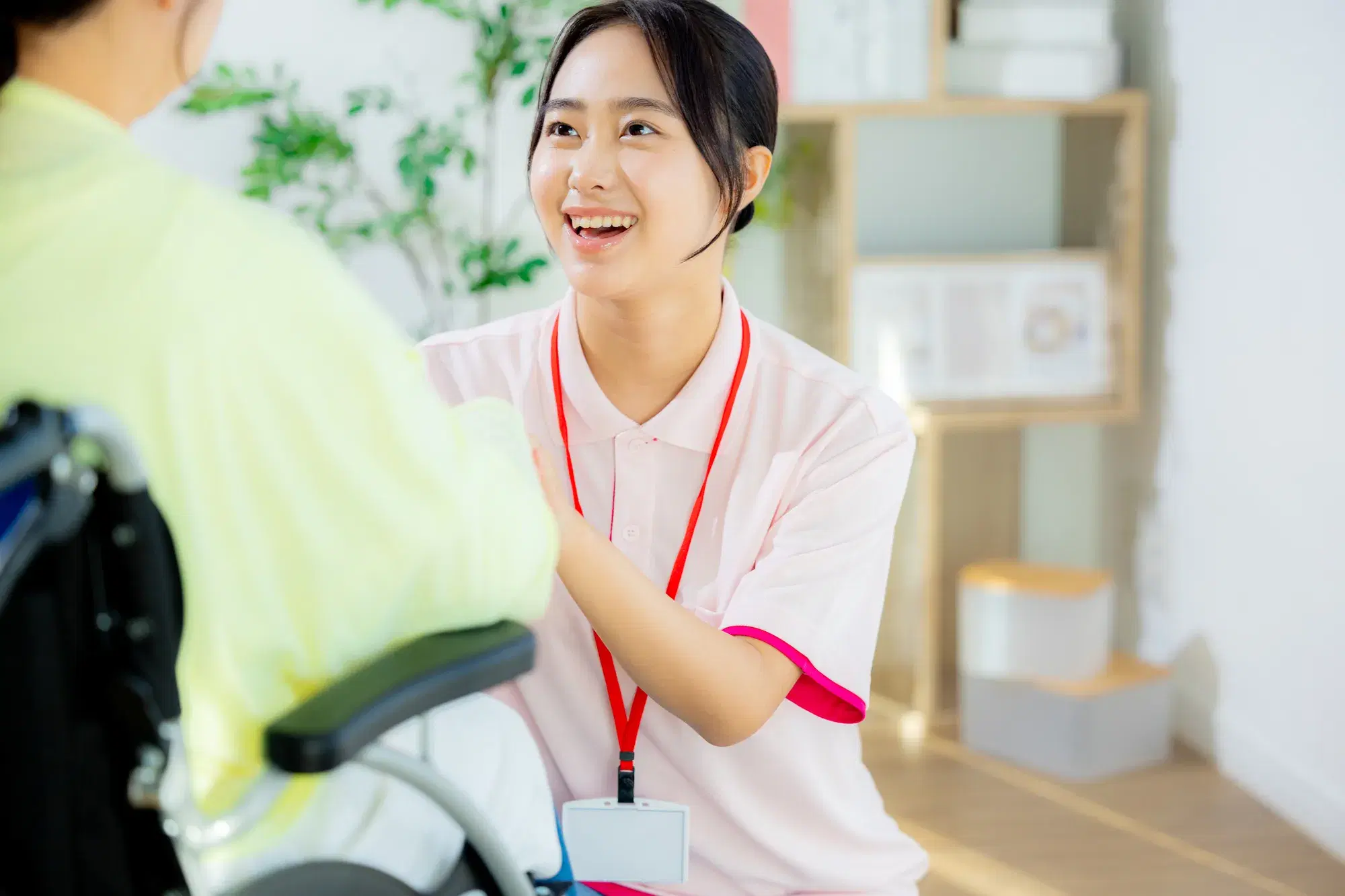 車椅子に座る黄色の服を着た人間と介護をしている首から赤いストラップを下げているピンク色の服を着た女性