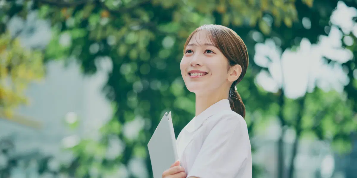 木を背景にバインダーを持ち笑顔を向ける職員女性