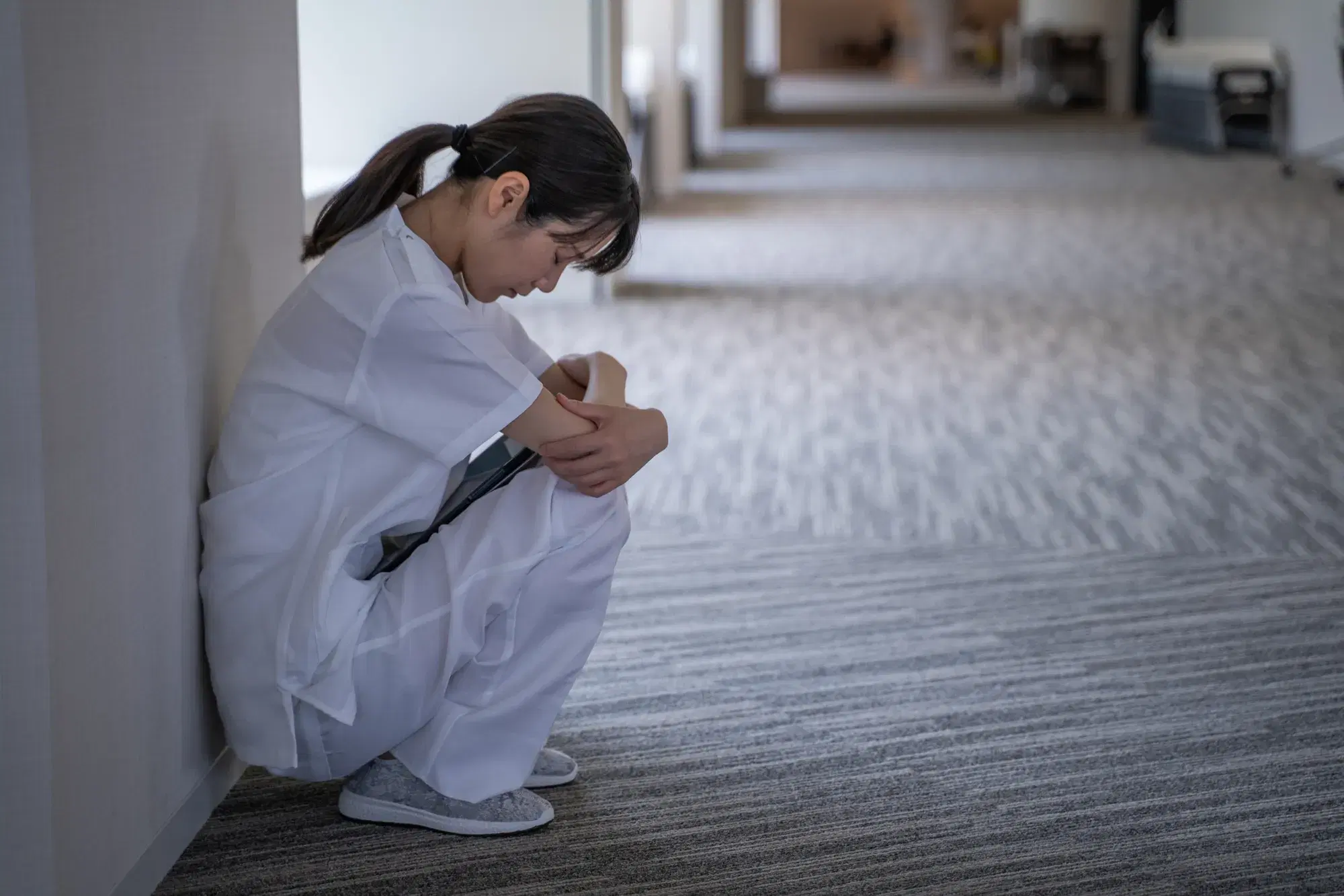 白い服を着た女性の医療従事者が廊下の壁際でしゃがみ込んでいる。