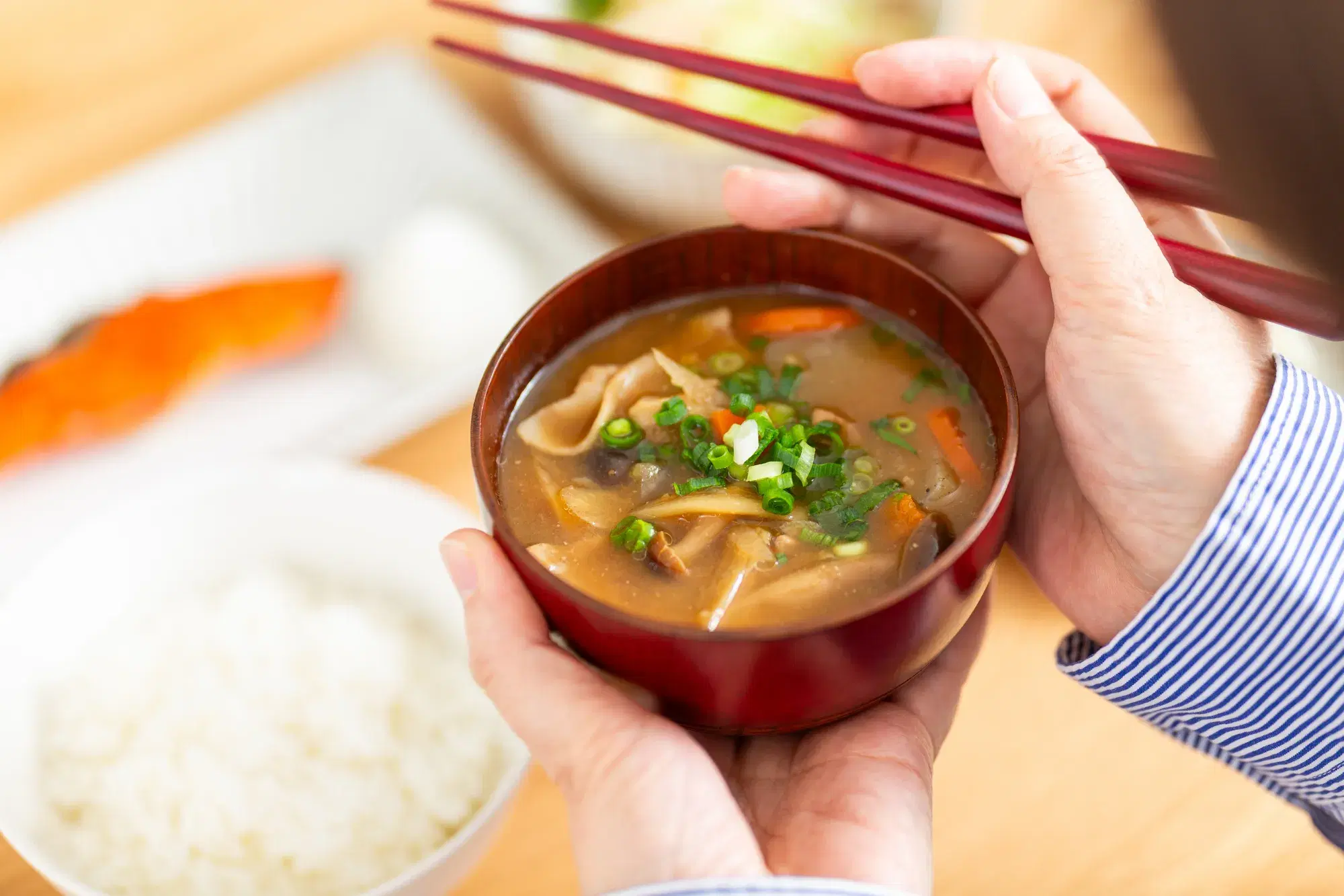 青い服を着た人が鮭とごはんとみそ汁を食べており、手には箸とみそ汁を持っている。