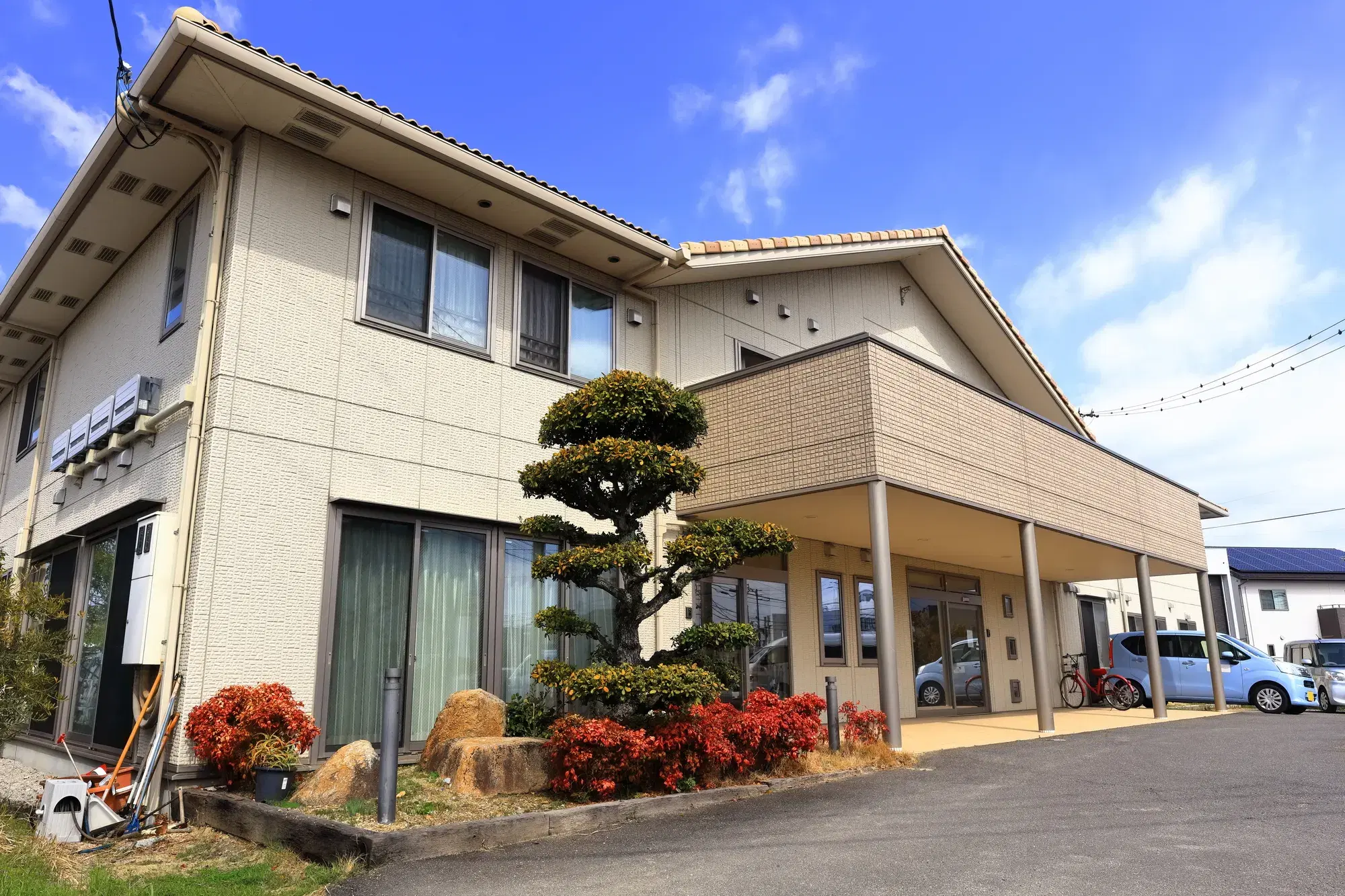 正面に大きな緑の木と低い位置に赤い植物が飾られている大きな一軒家