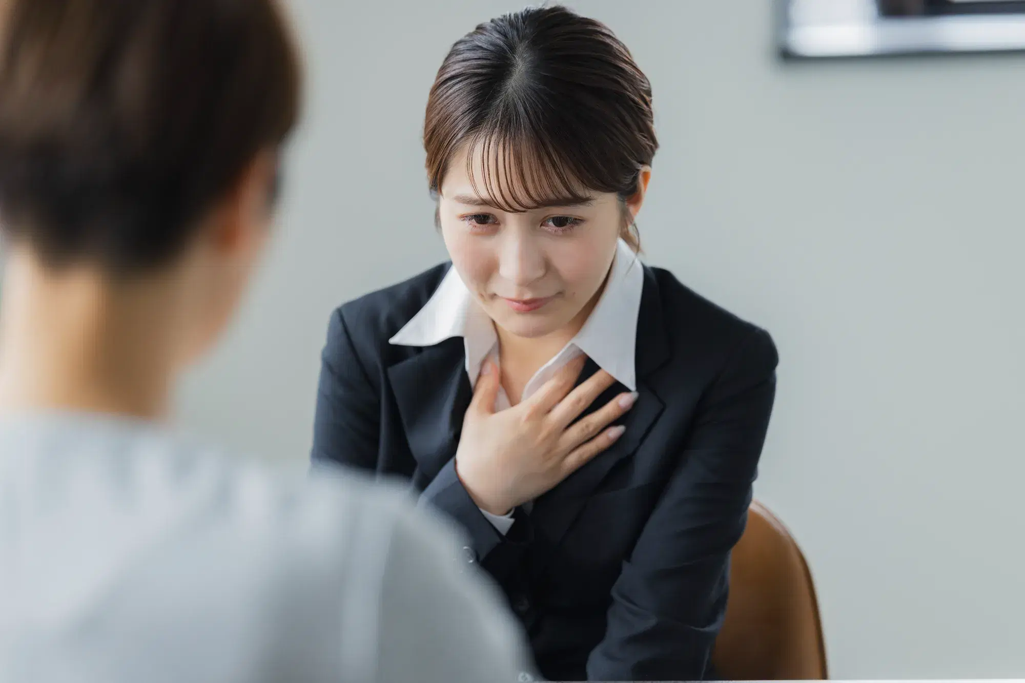 手前にグレーの服を着た人物がおり、奥で黒いスーツを着た女性が胸の上に手を置いている。