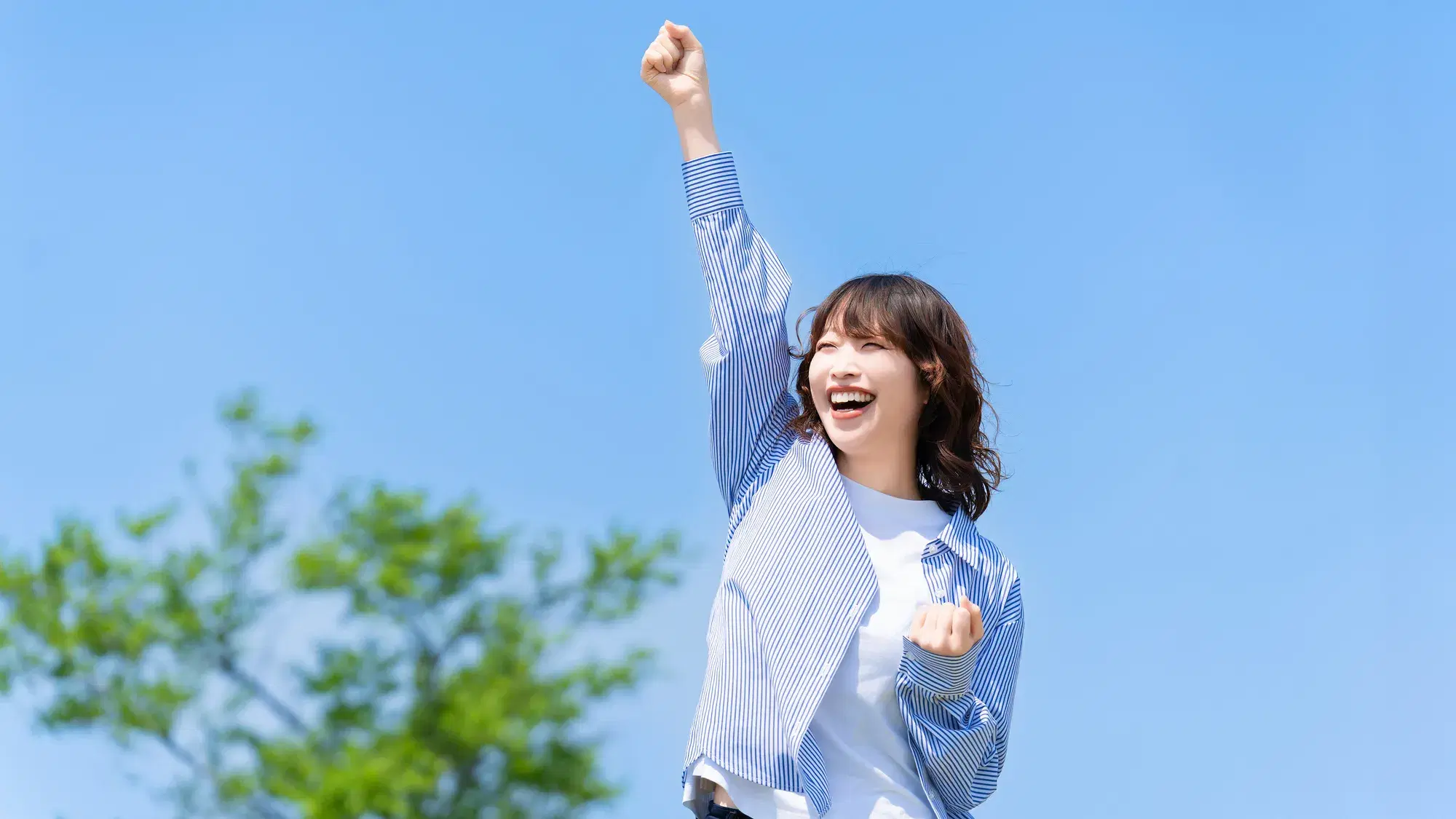 青空の下で笑顔で拳を上に突き上げる青い服をきた女性
