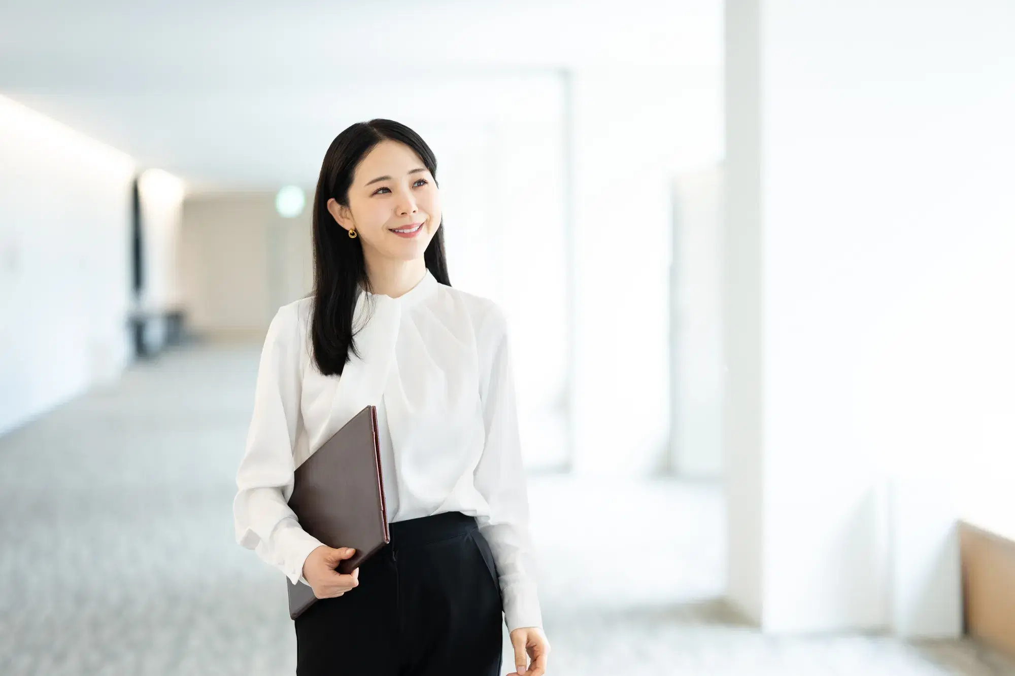 右腕に茶色のバインダーを抱えて廊下を歩く白い服を着た女性