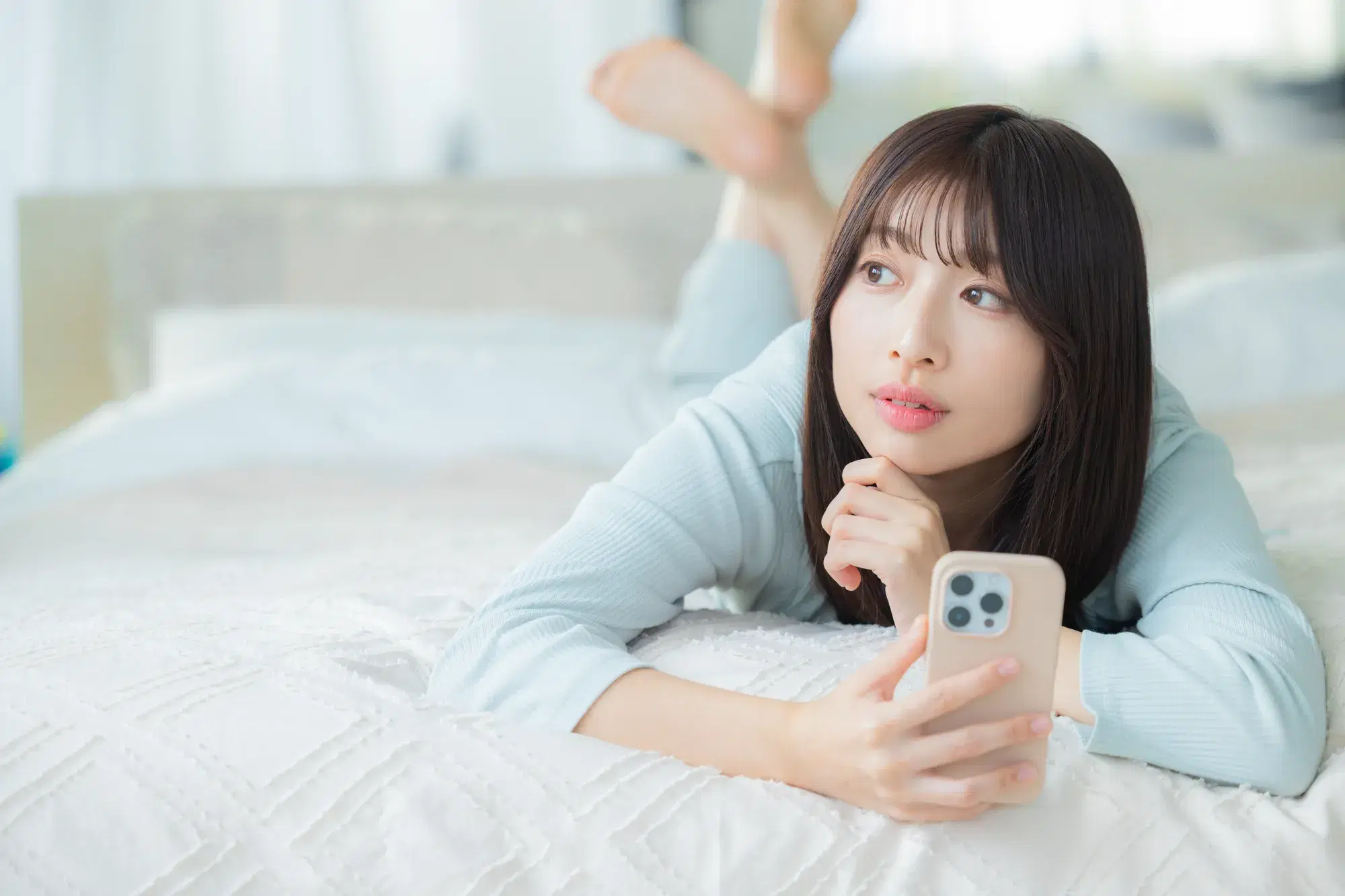 水色の服を着た女性がベットの上で寝ころびながらスマホを右手で持ち外を見る