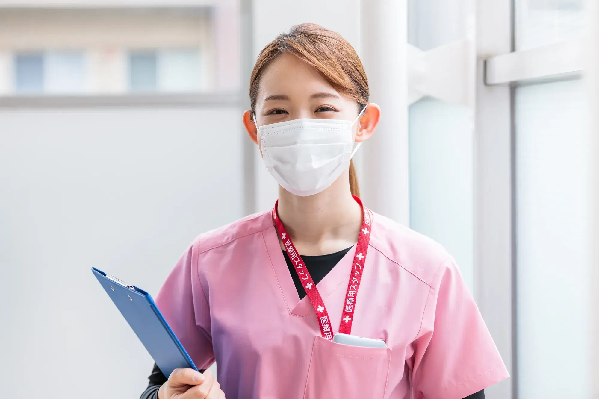 ピンクの医療用の服を着たマスクをした女性がバインダーを持ちながら笑っている