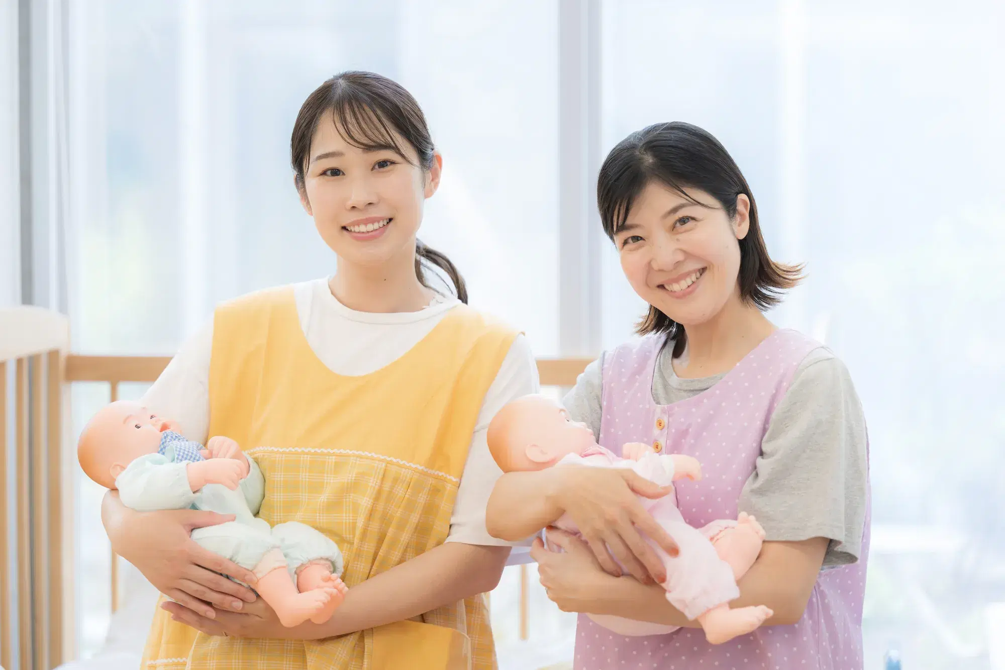 白い服と黄色のエプロンを着た女性と、グレーの服と紫色のエプロンを着た女性が、それぞれ赤ちゃんの人形を抱いて立っている。