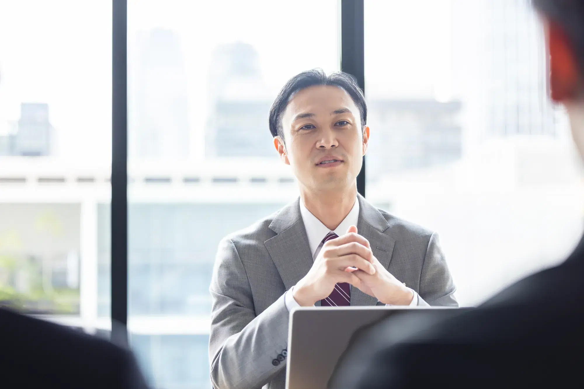 手前に黒いスーツを着た人物たちがおり、奥でグレーのスーツを着た男性が手を組んで座っている。