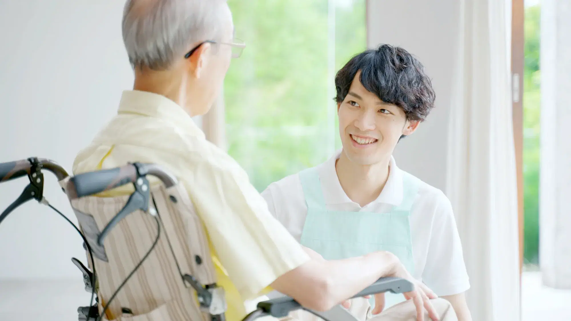 黄色の服を着て車いすに座っている男性と白い服に水色のエプロンをした介護士が話をしている。