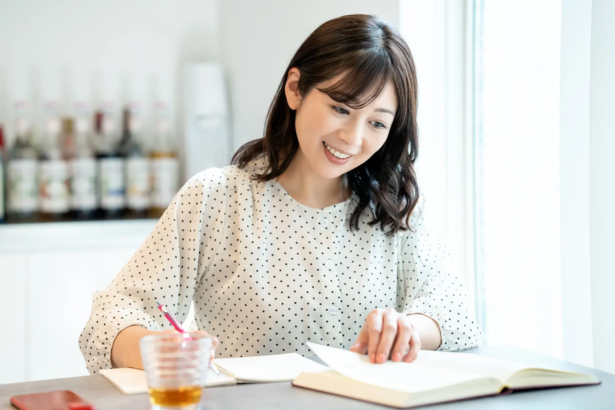 白い生地に黒の小さいドット柄の服を着た女性が本をっめくりながら机に麦茶を置いて勉強している
