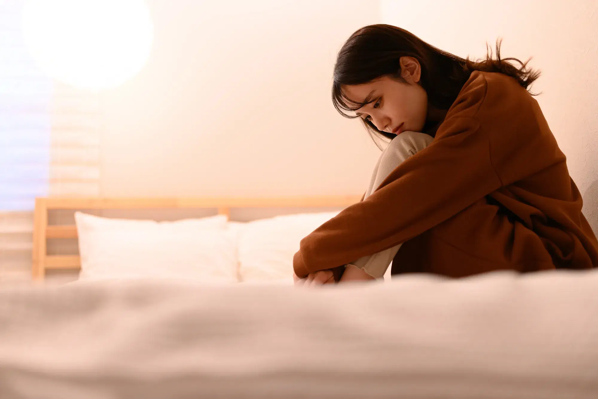 茶色い服を着た女性がベッドの上で体育座りをしている