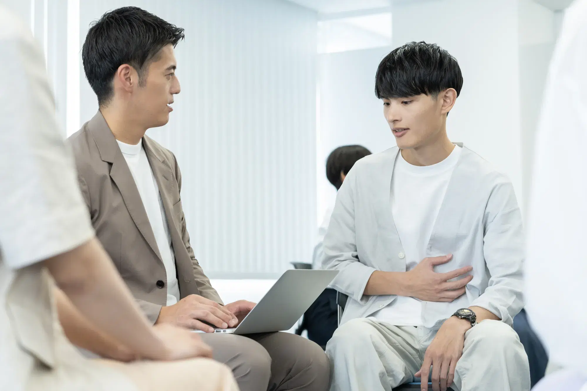 腹部を手でおさえる男性と向かいに座りPCを操作する男性