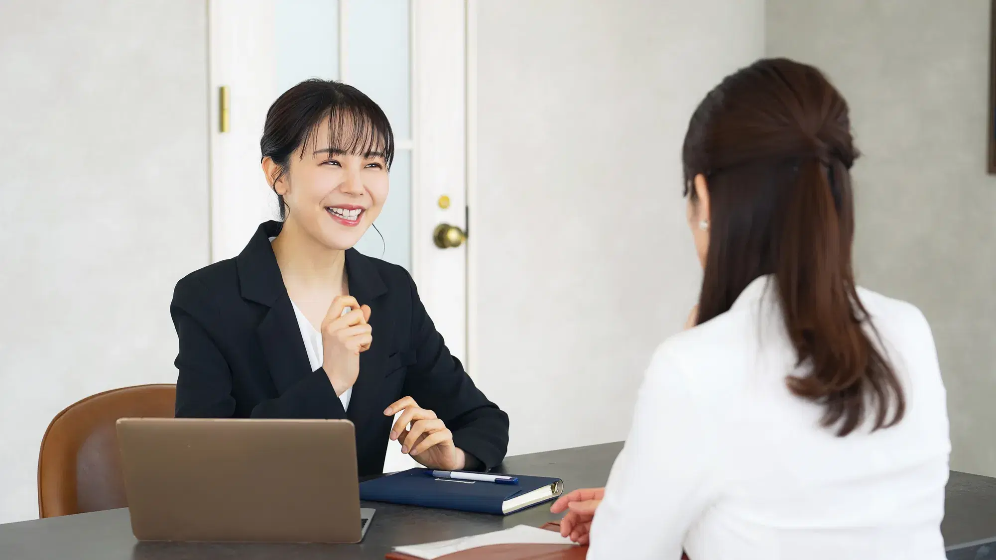 黒いスーツを着ており髪の毛を一つに結んだ女性が白い服を着たハーフアップの女性と部屋で話をしている。