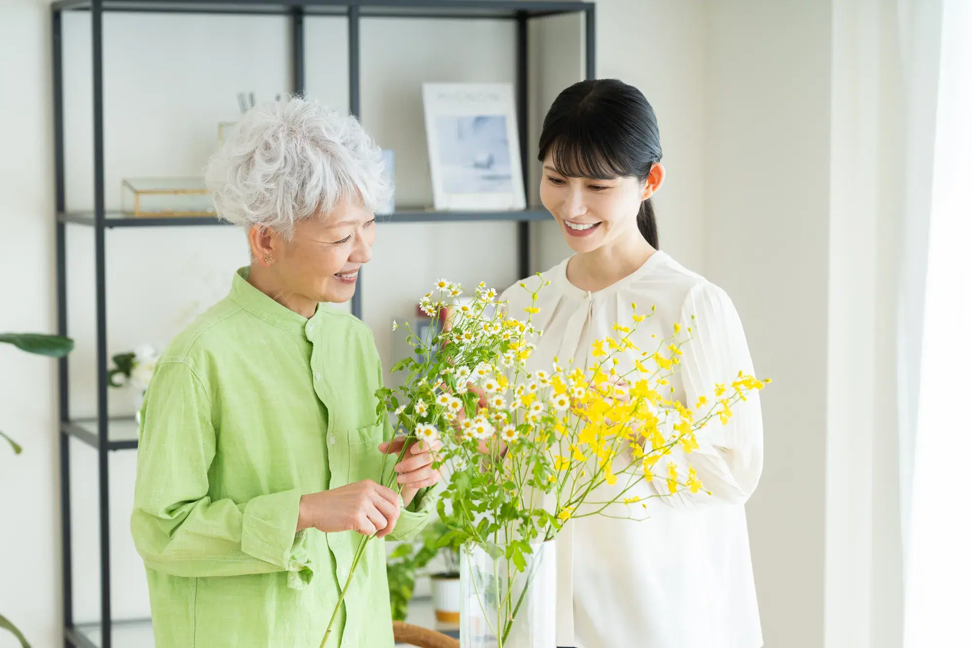 緑色のシャツを着た女性と白い服を着た女性が、花瓶に黄色の小花を生けている。
