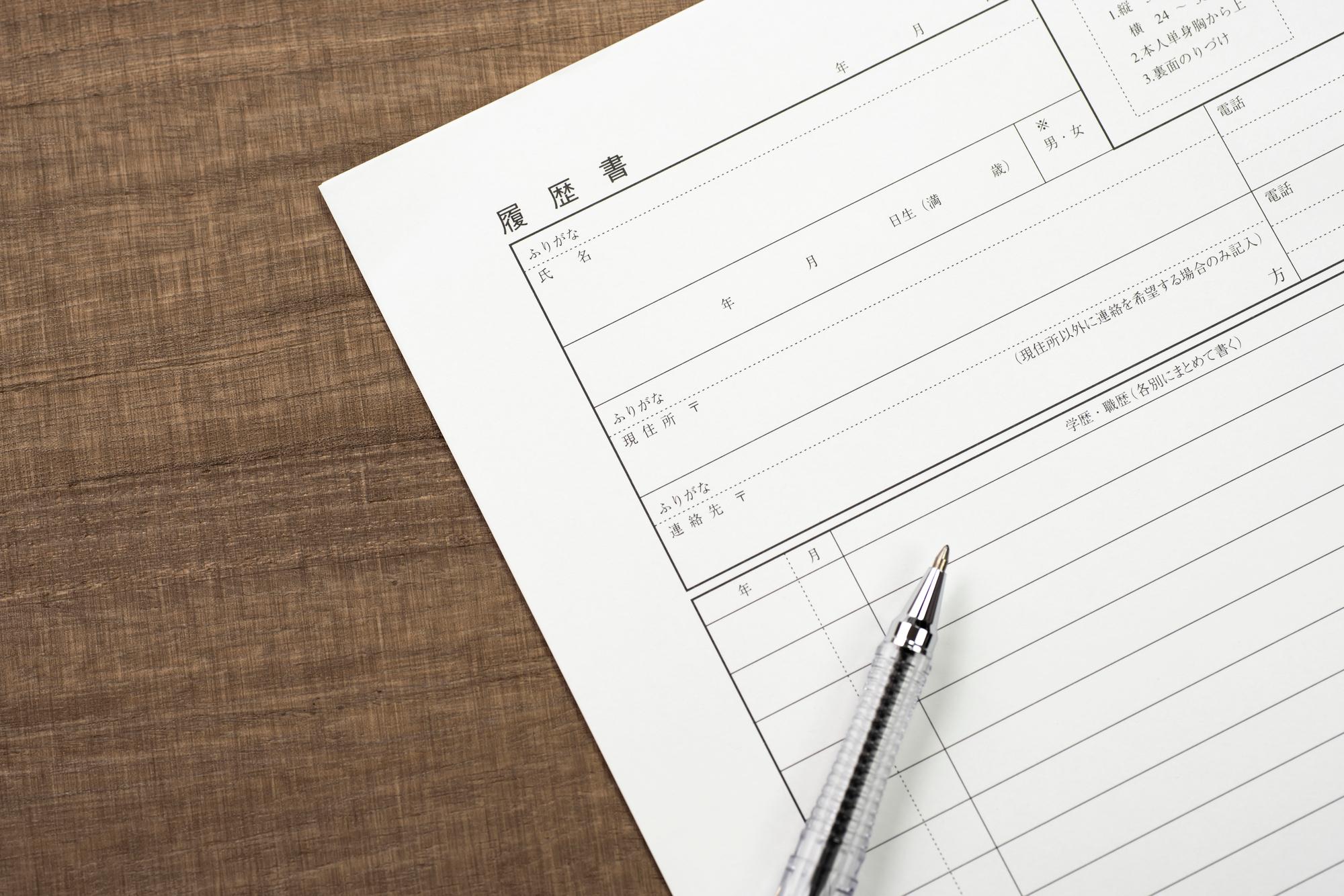 木目のテーブルの上に「履歴書」と書かれた用紙と銀色のペンが置かれている様子が写っている。