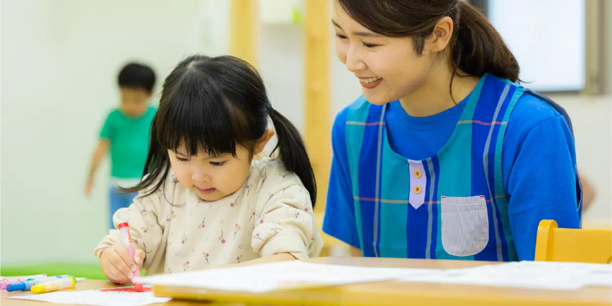クレヨンで絵を描く幼児を見守る職員の女性