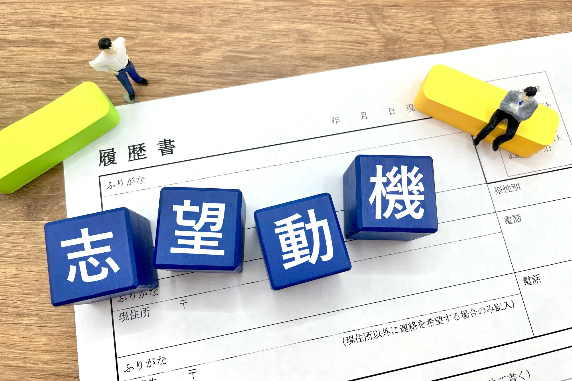 「履歴書」と書かれた用紙の上に、「志望動機」と書かれた青色のキューブ型ブロック、緑色と黄色のブロック、および2体のミニチュア人形が置かれている様子が写っている。