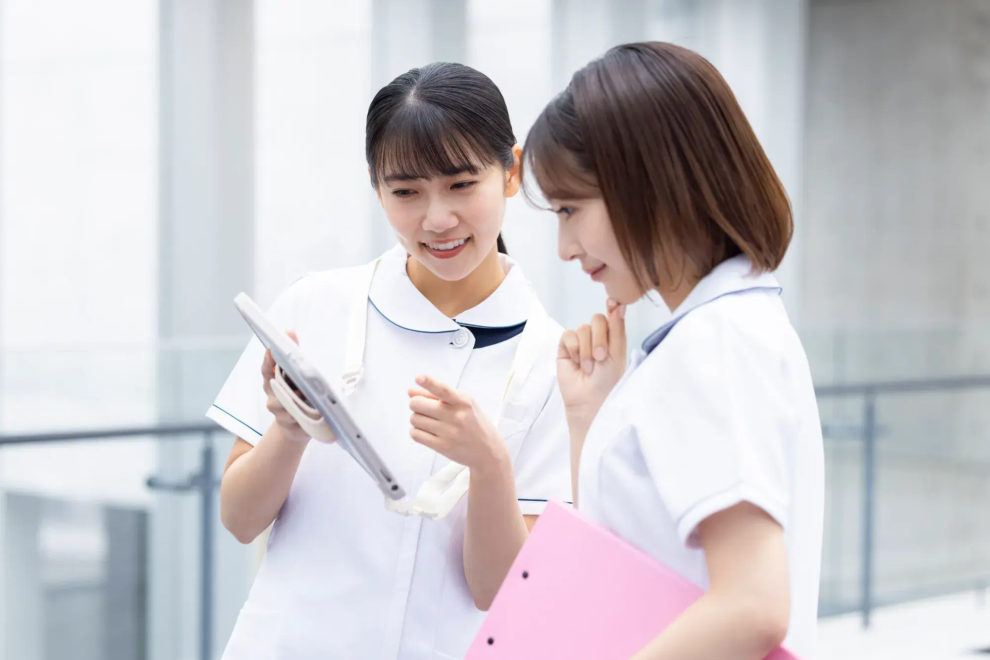 看護婦二人がタブレットを見て話し合っており、一人はピンクのバインダーを持っている。