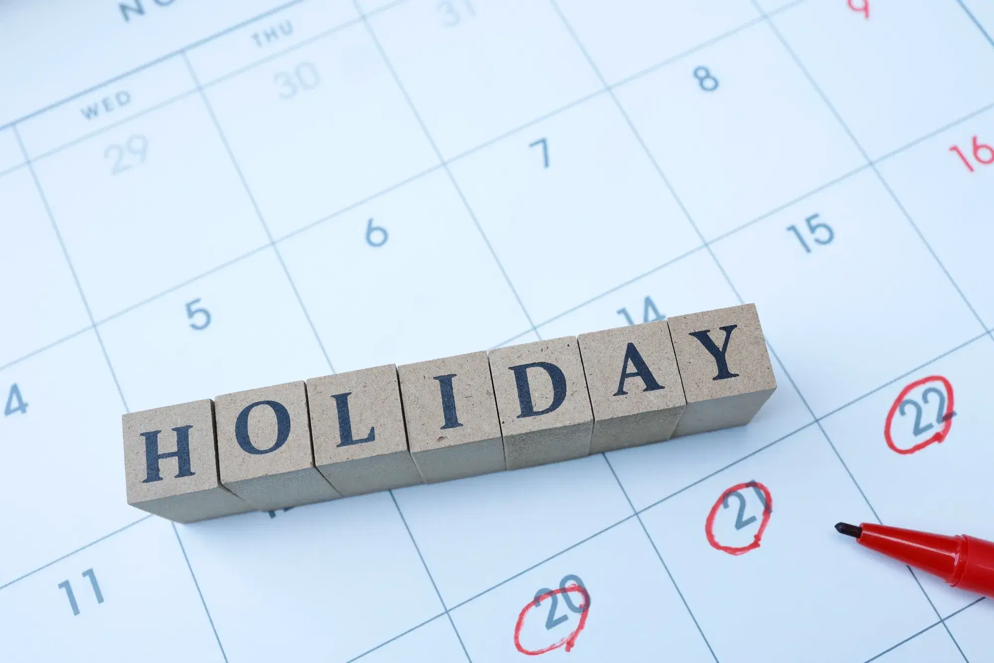 日付に赤丸がつけられたカレンダーの上に、「HOLIDAY」と書かれた木製ブロックと赤色のペンが置かれている様子が写っている。