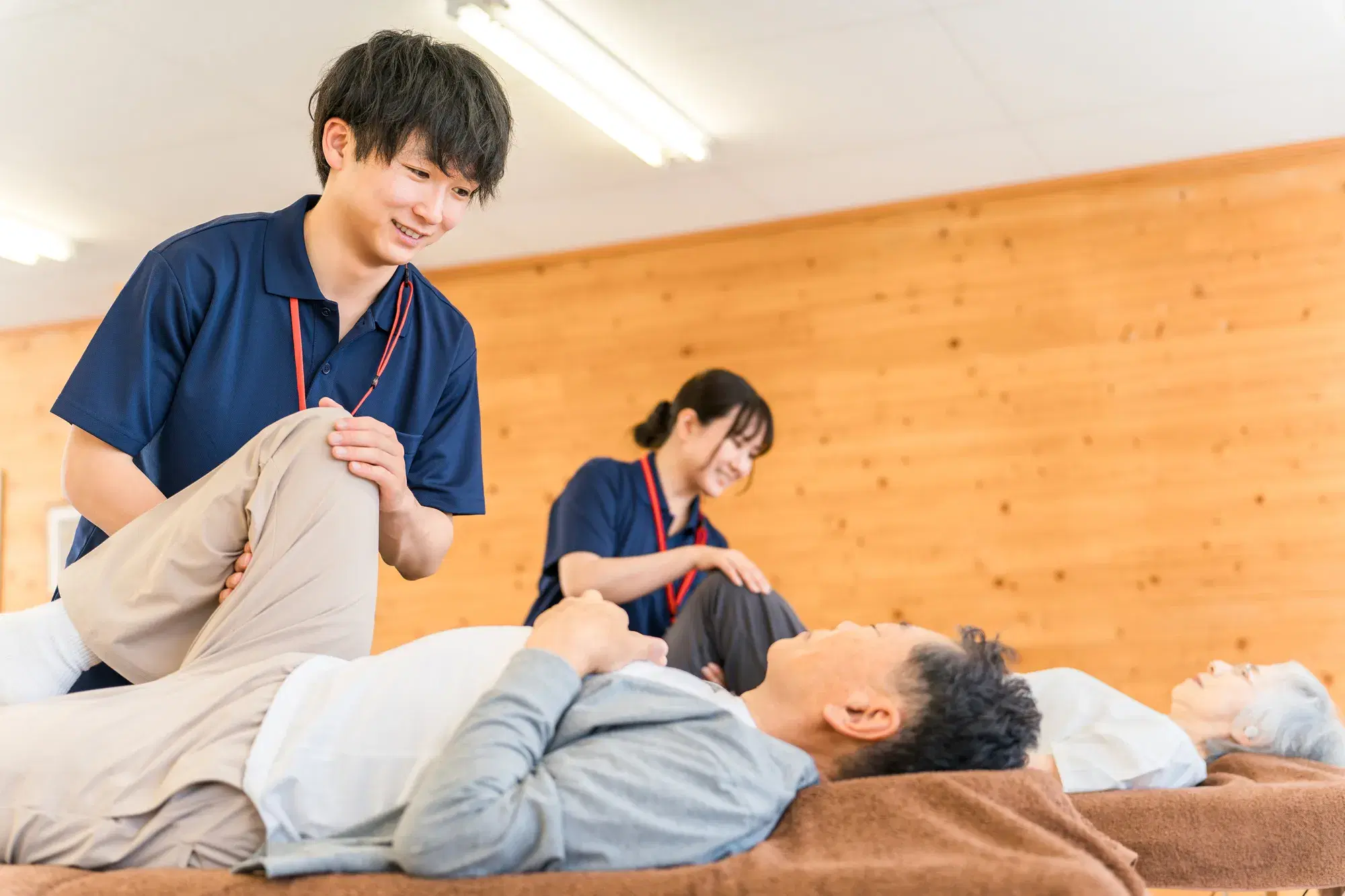 紺色の服に赤色のストラップをつけている男性がグレーの服にベージュのズボンを履いた男性に施術をしている