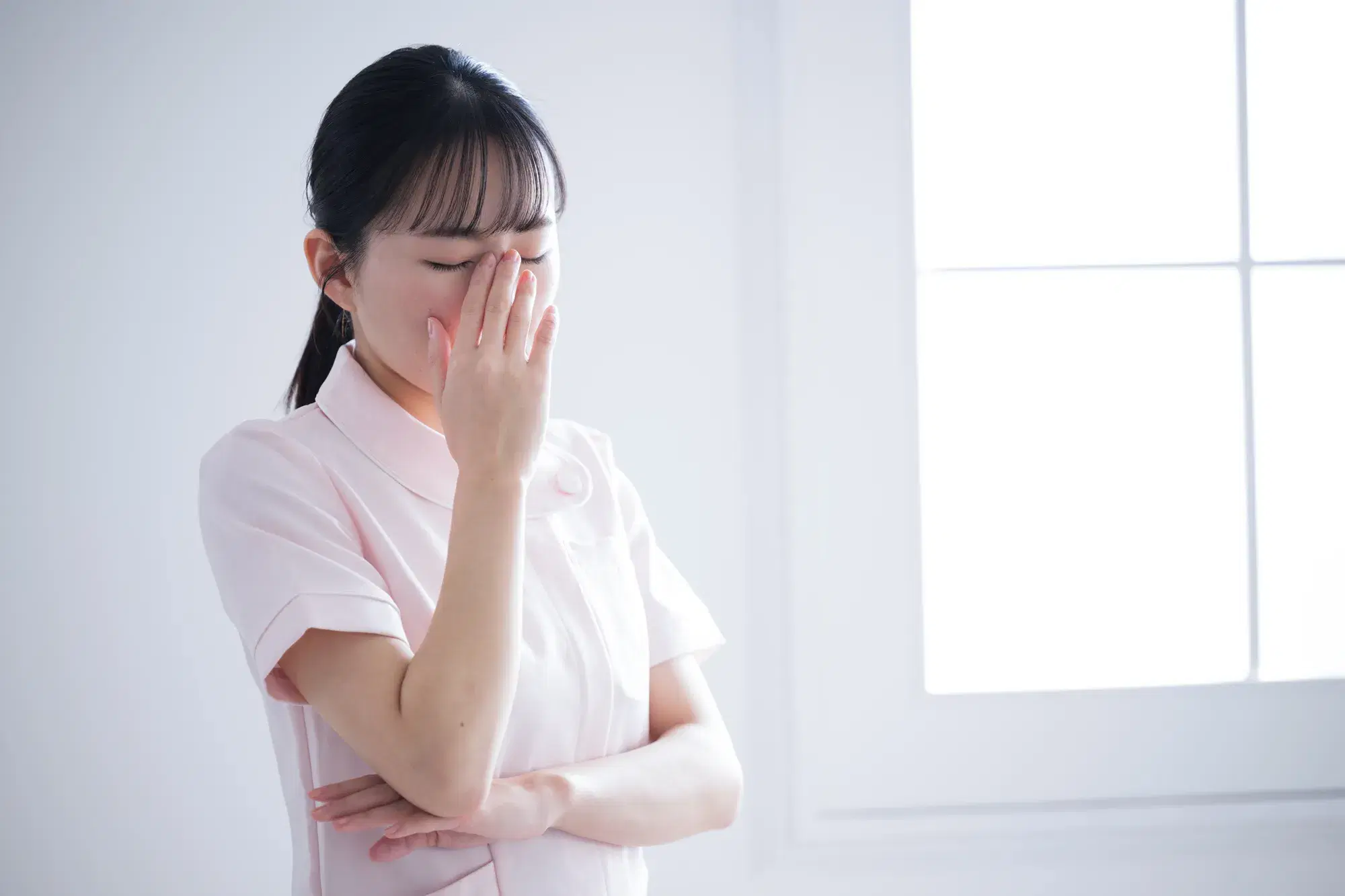 薄いピンクの医療用の服を着た女性が目と目の間に手を当てており困っている様子である