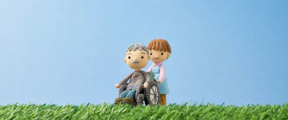 芝生の上に車椅子に座る年配の男性人形と水色のエプロンを着用した女性人形