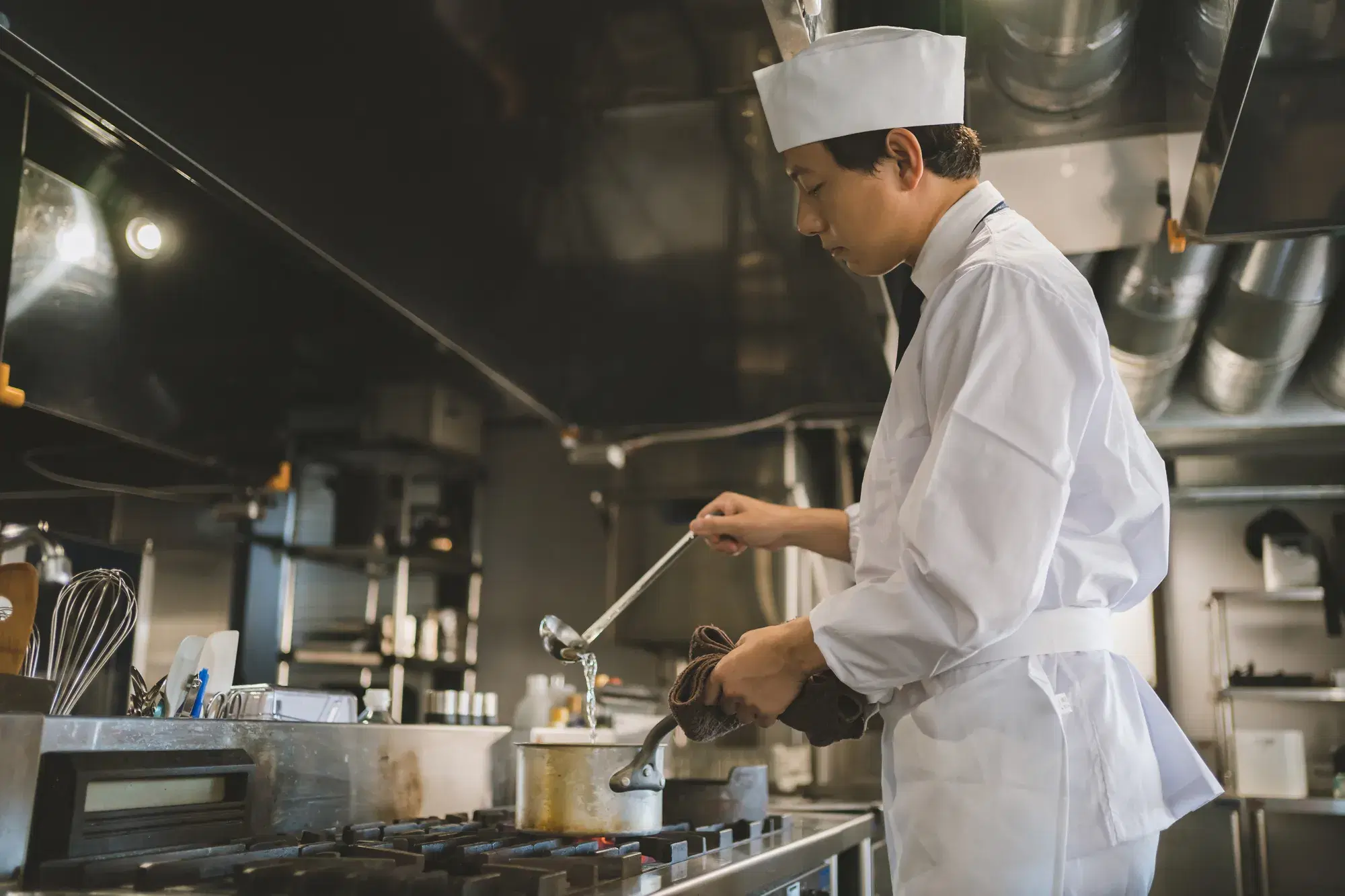 片手に鍋、片手におたまを持った白い作業服を着た男性の料理人