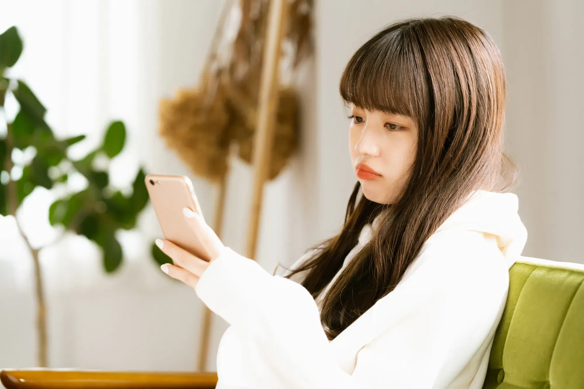 背もたれが緑のソファーに座っている片手にスマホを持った白いパーカーを着た女性