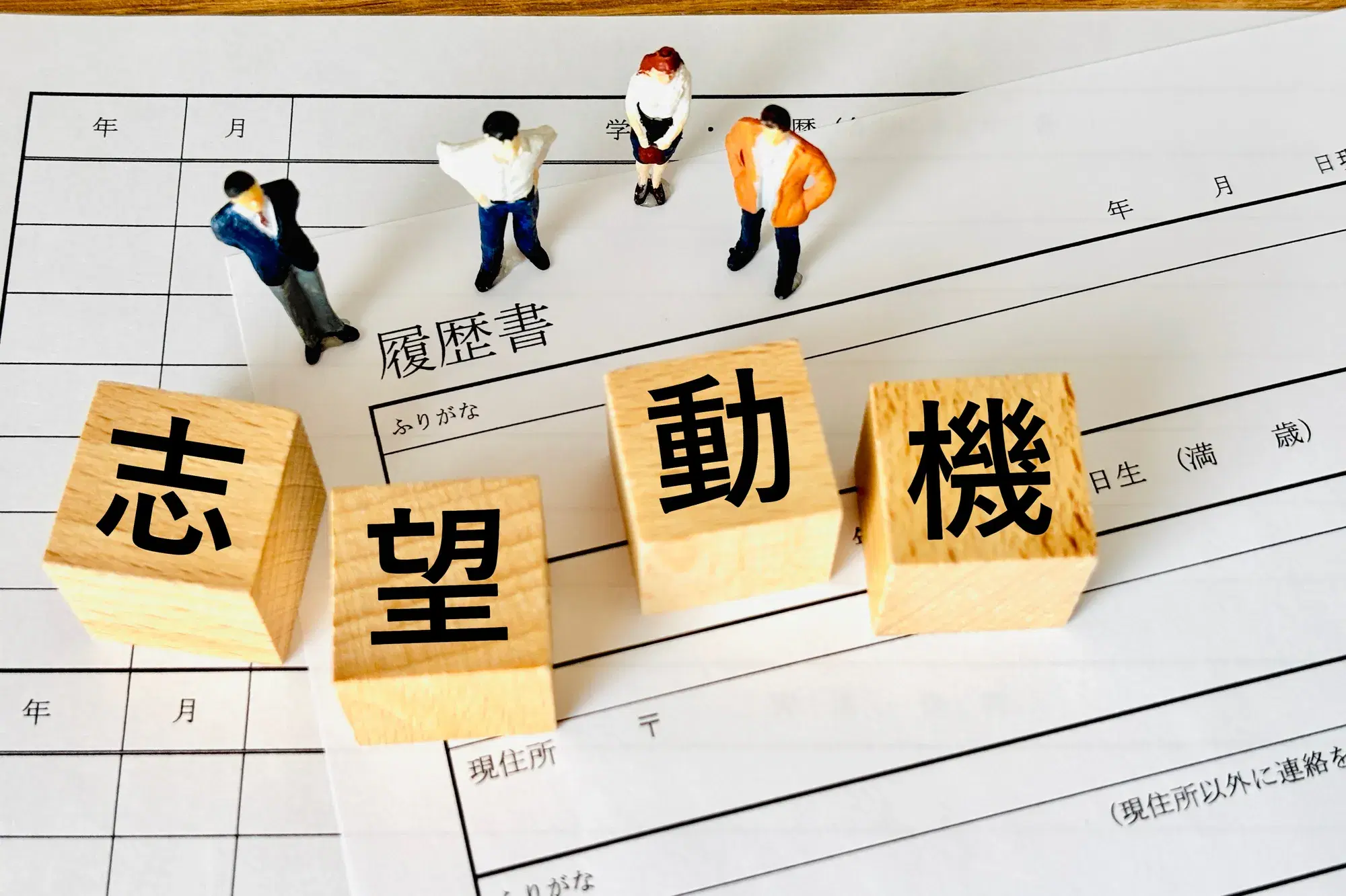 「履歴書」と書かれた用紙の上に「志望動機」と書かれた木製ブロックと、複数のミニチュア人形が置かれている様子が写っている。