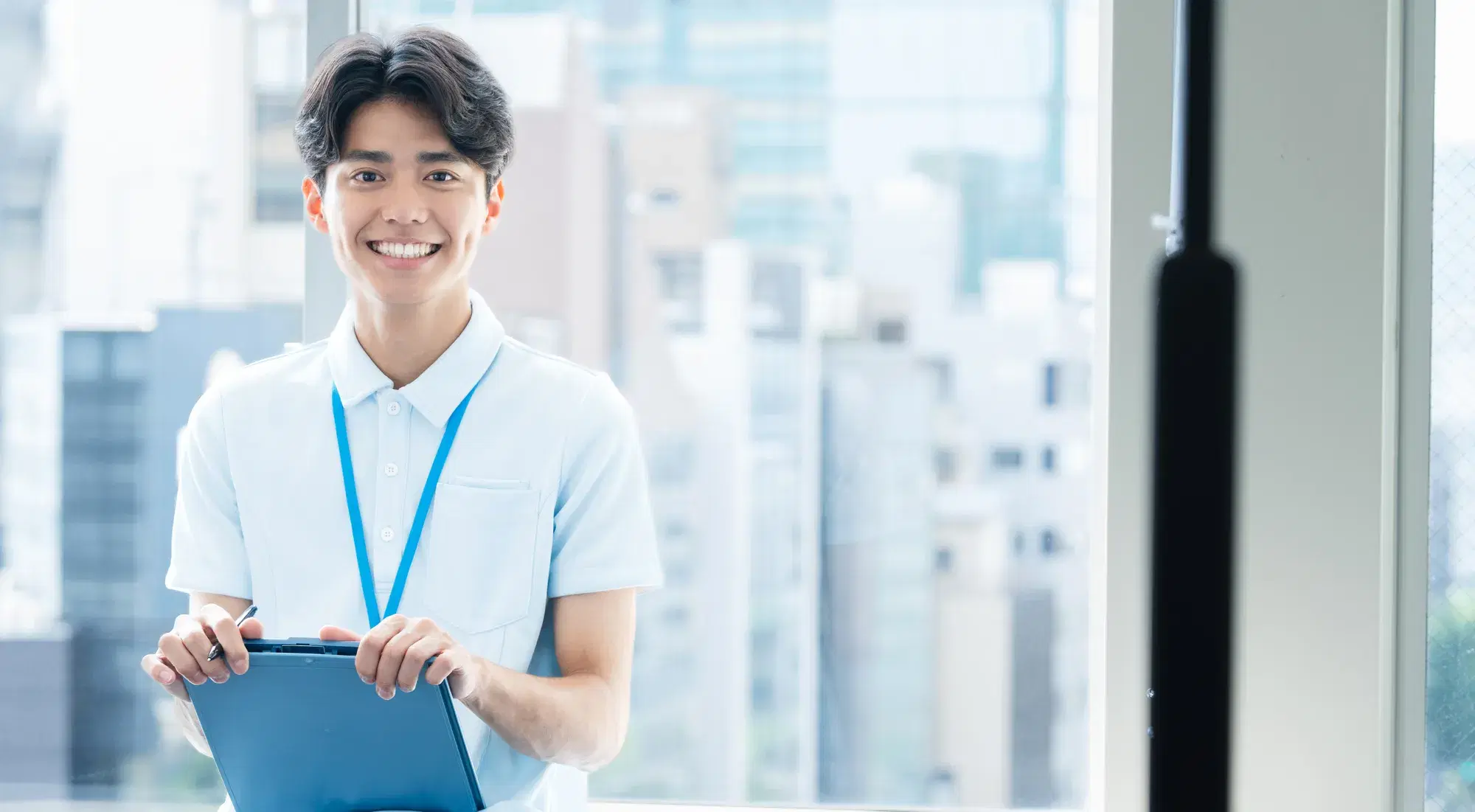 大きな窓の前に若い男性が青いバインダーを両手に持ち笑顔で立っている