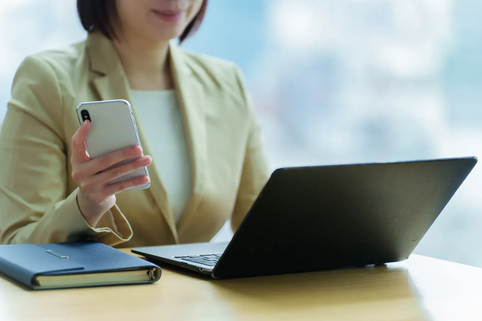 白い服を着た女性が右手にスマホを持って机のPCを見ている