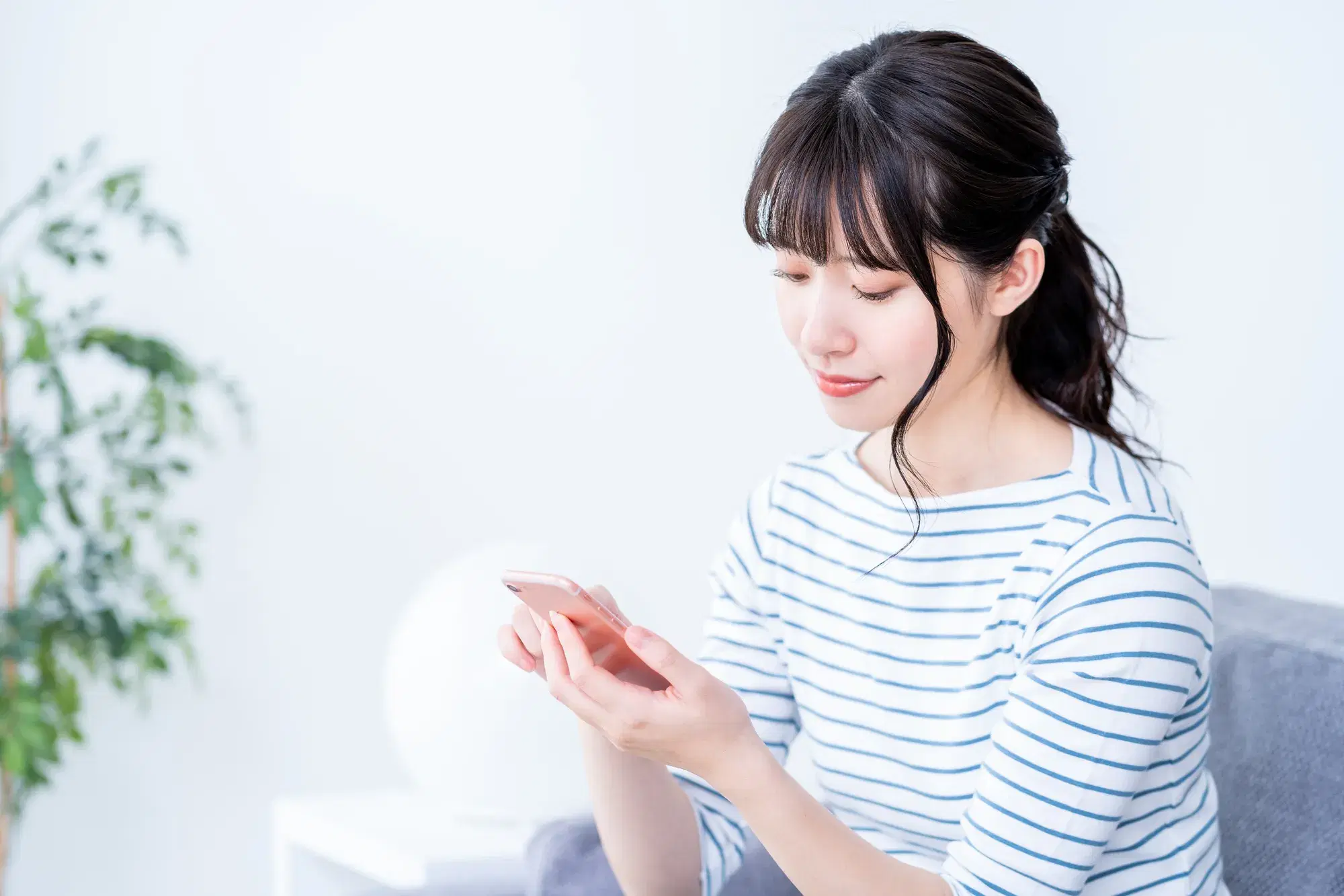 ボーダーの服を着た女性が灰色のソファに座ってスマホをみている。